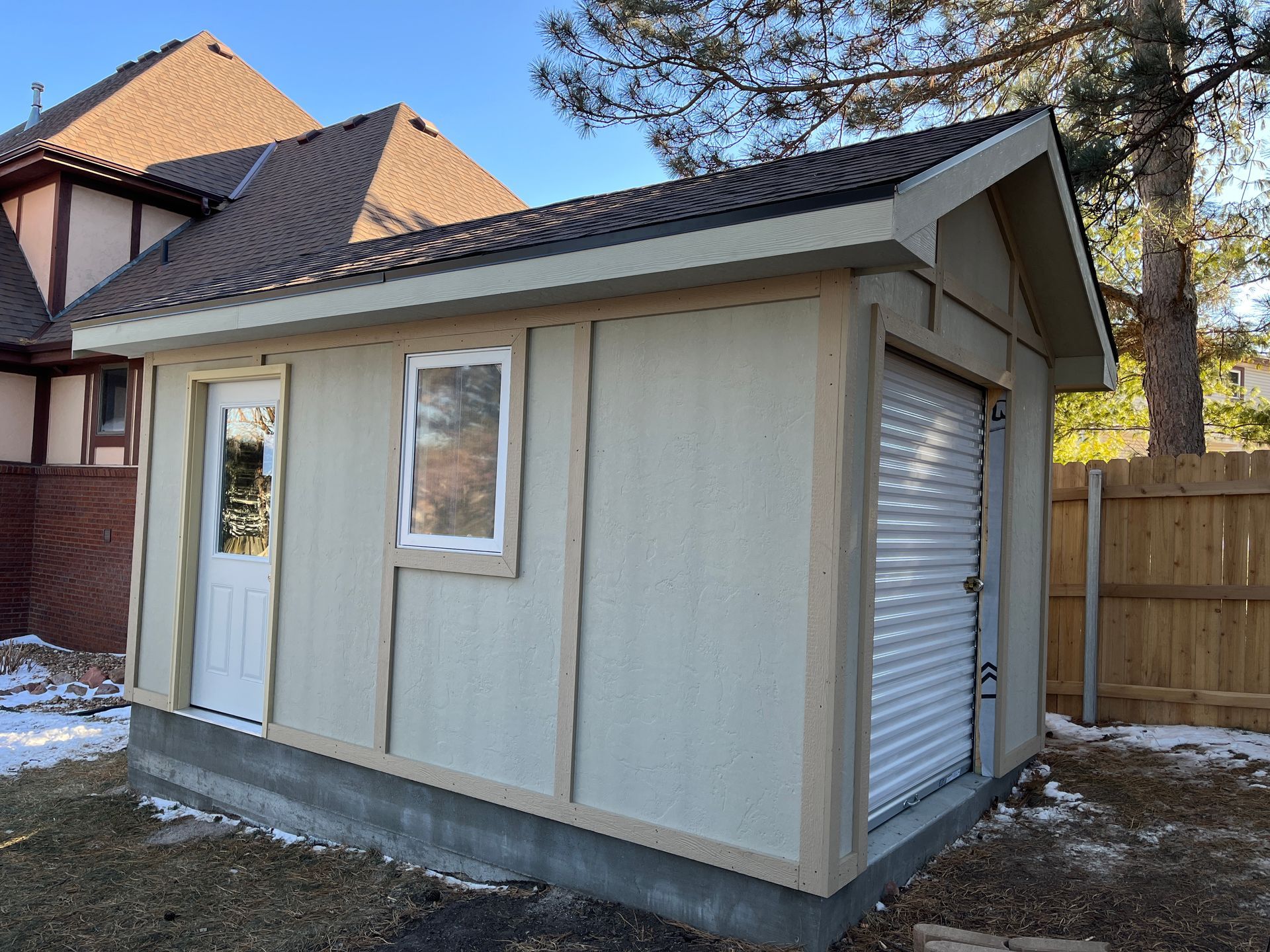 Small, light-colored shed with a roll-up door, window, and door. Brown roof, beige siding, and a concrete foundation.
