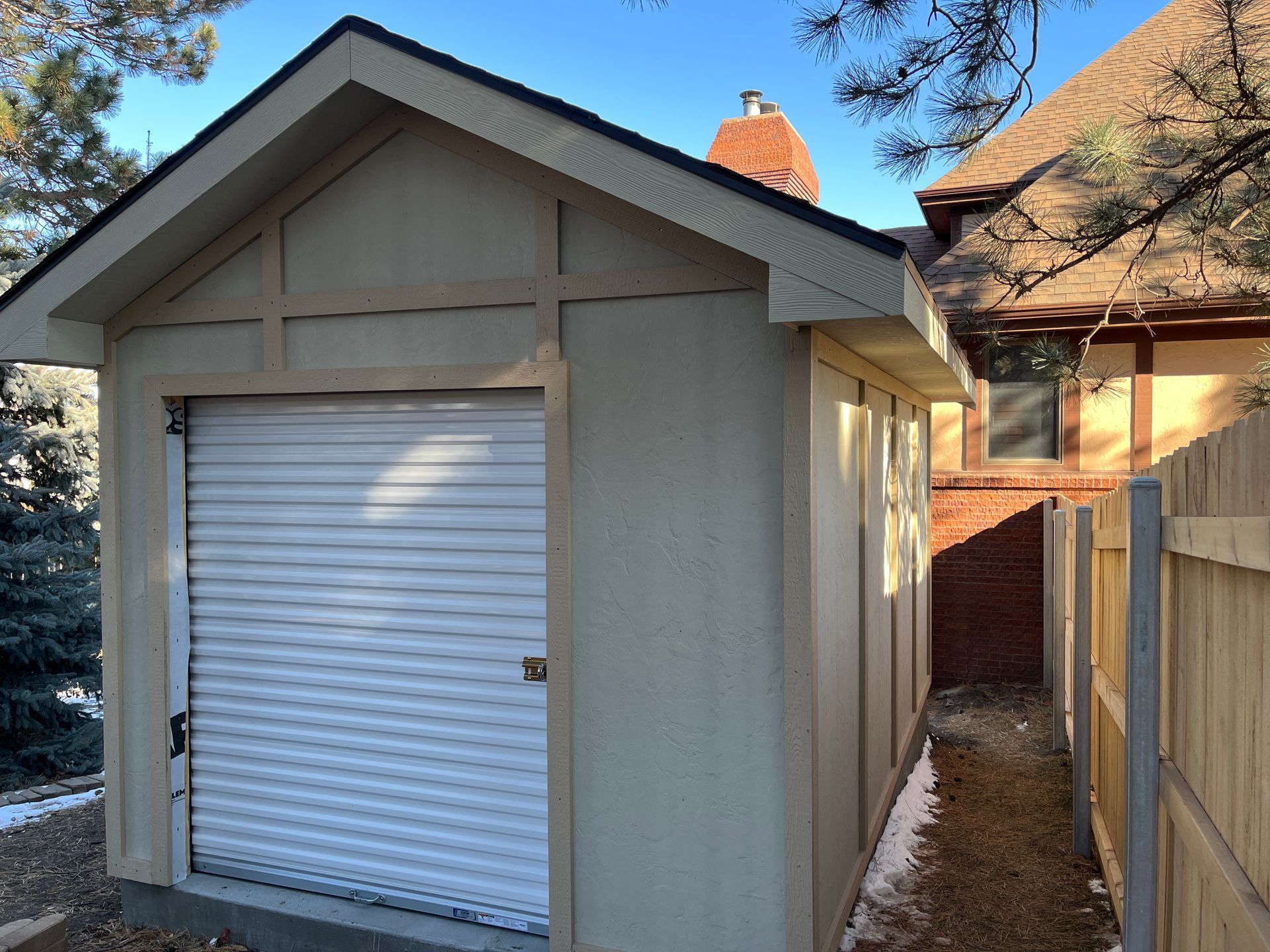 Tan shed with a white roll-up door, nestled next to a house. A narrow pathway runs along the side.