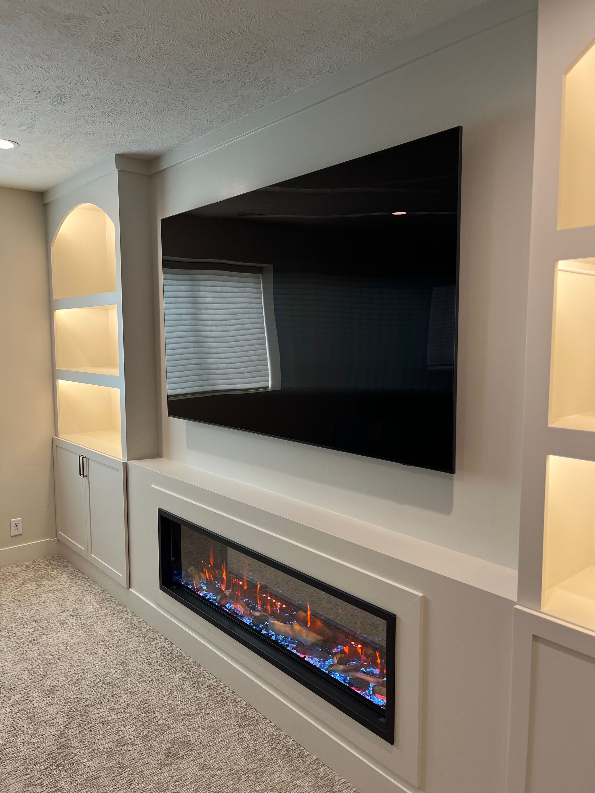 Built-in entertainment unit with TV above a fireplace, flanked by lit shelves, set against a neutral-toned wall and carpet.