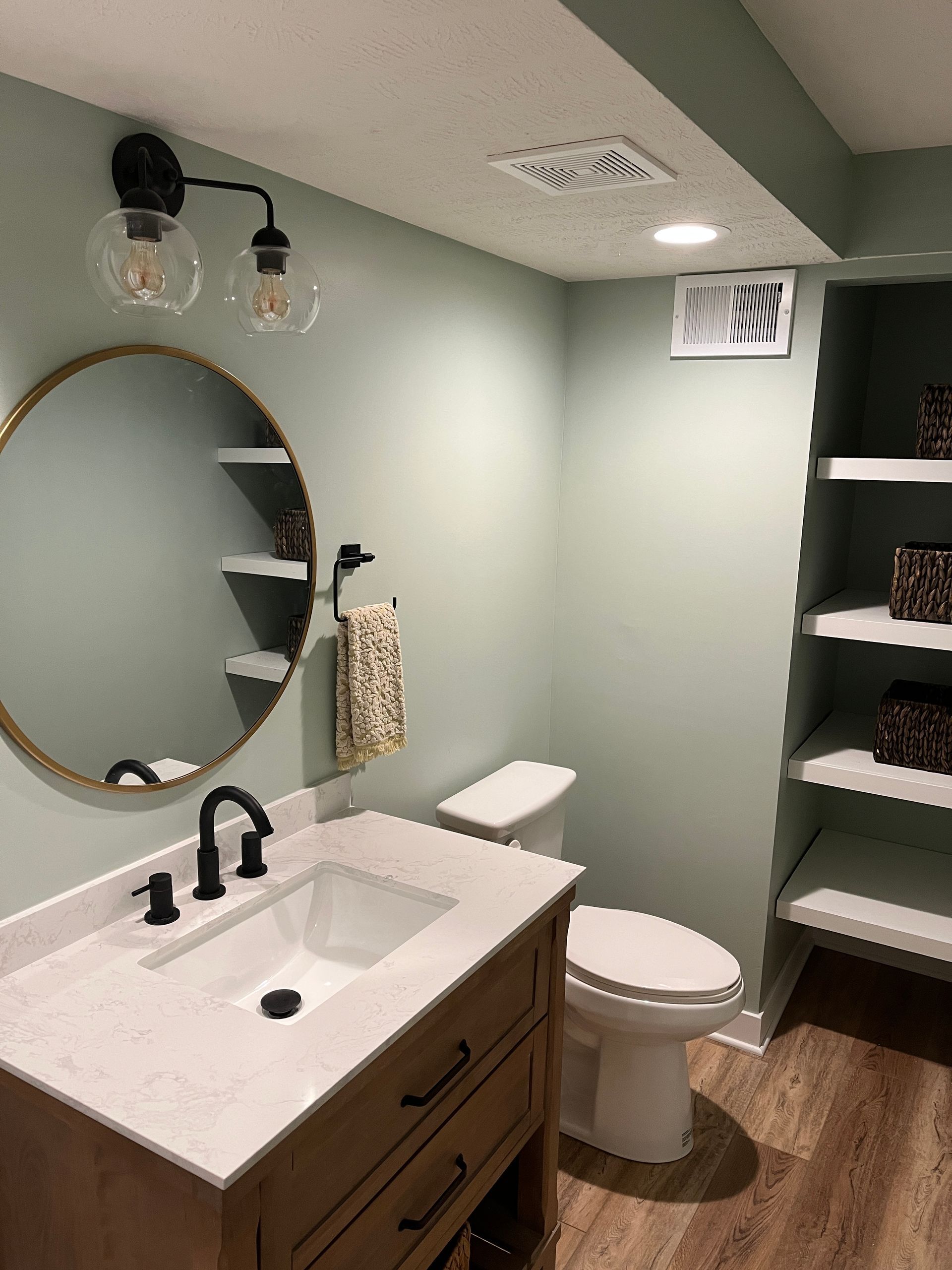 A bathroom with a sink , toilet , mirror and shelves.