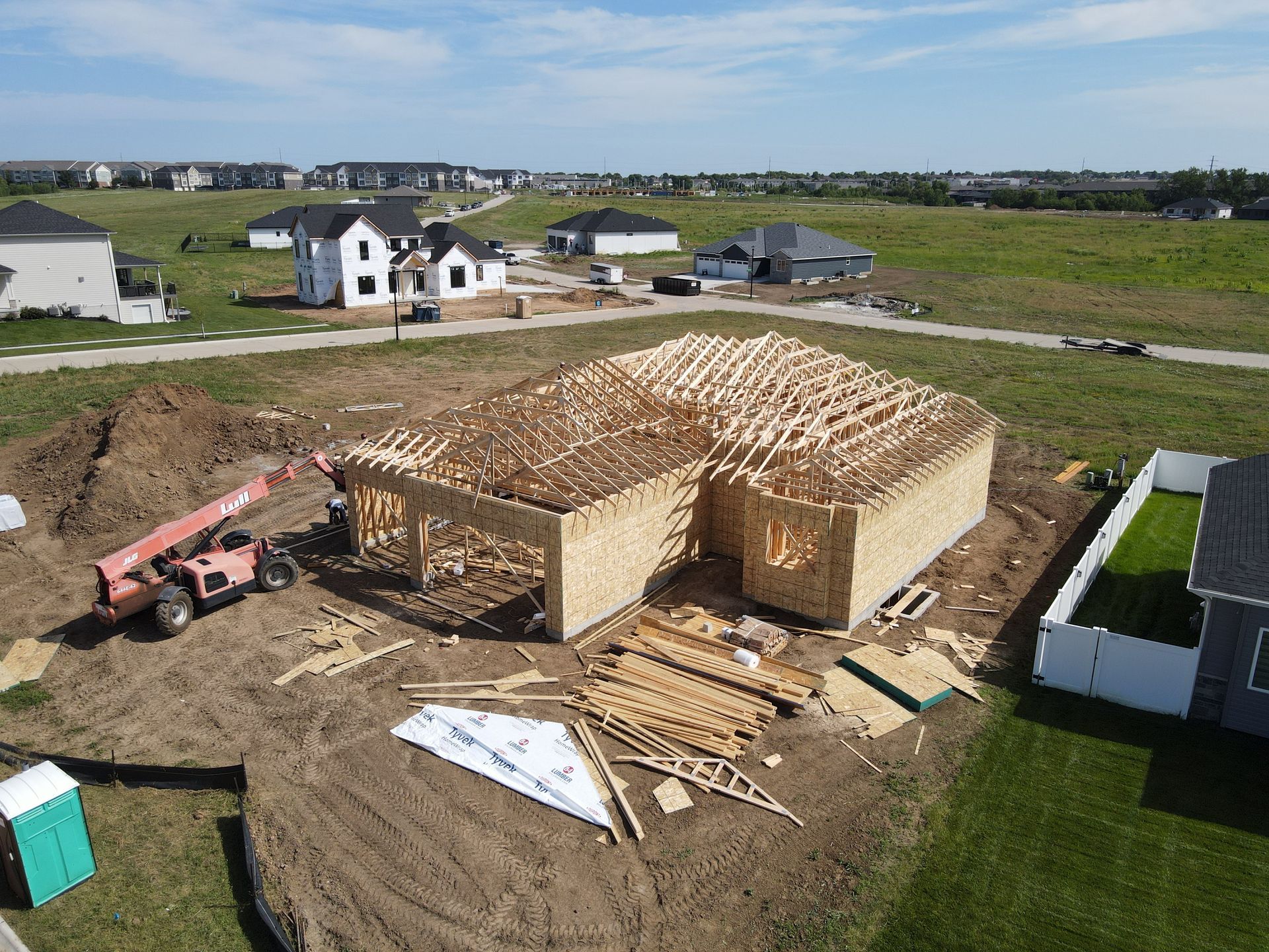 Construction site with a house frame under construction. A pink lift, building materials, and other homes are visible.