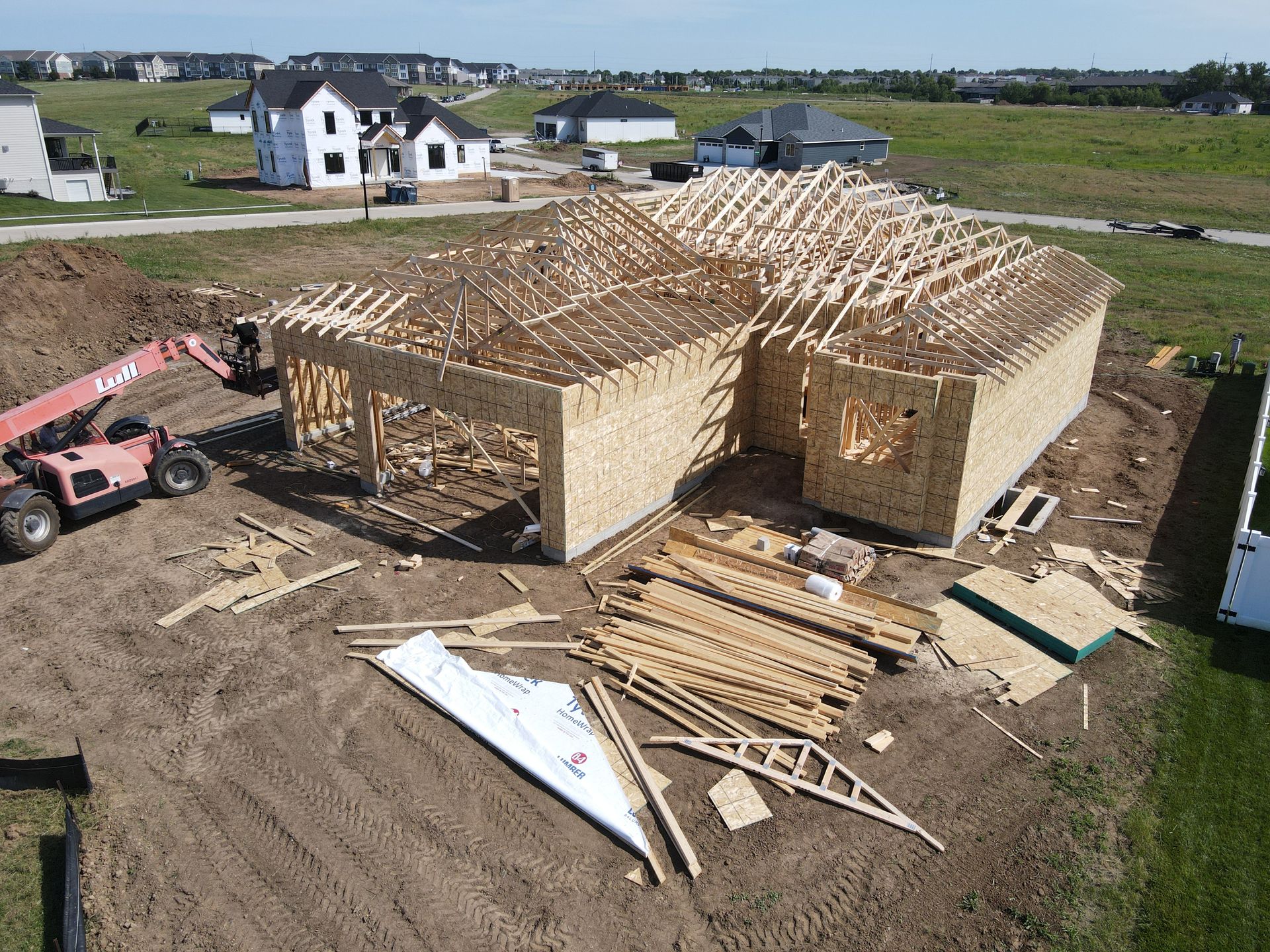 A house under construction; wooden frame visible. A crane and building materials sit on a dirt lot in a residential area.