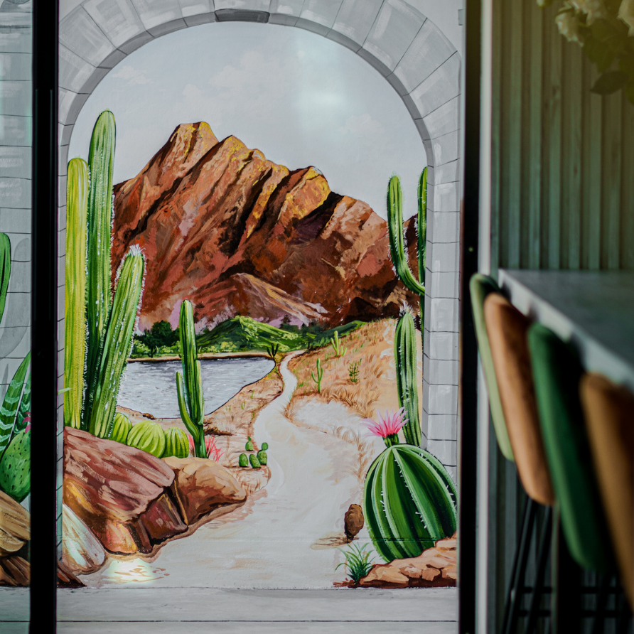 Desert landscape mural on glass panel: cacti, mountains, path, water, and arch design.