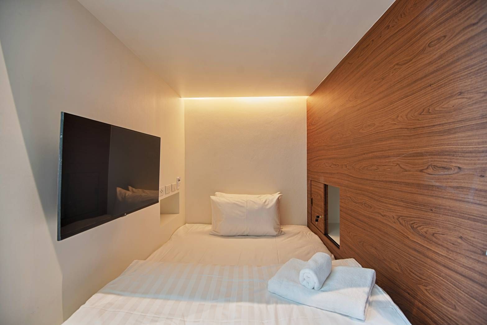 Small hotel room with a bed, TV, and wood-paneled wall. White linens, neutral colors, minimalist design.
