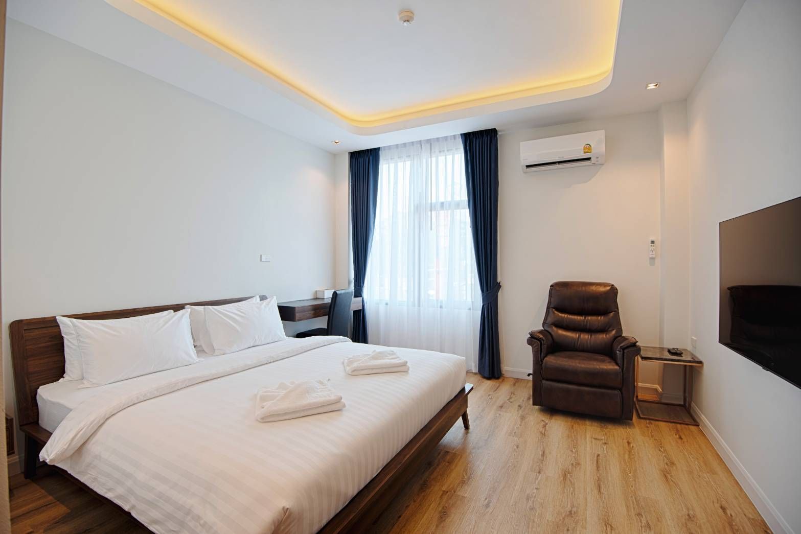 Hotel room with a bed, desk, armchair, and TV. Light wood floors, white walls, and blue curtains.