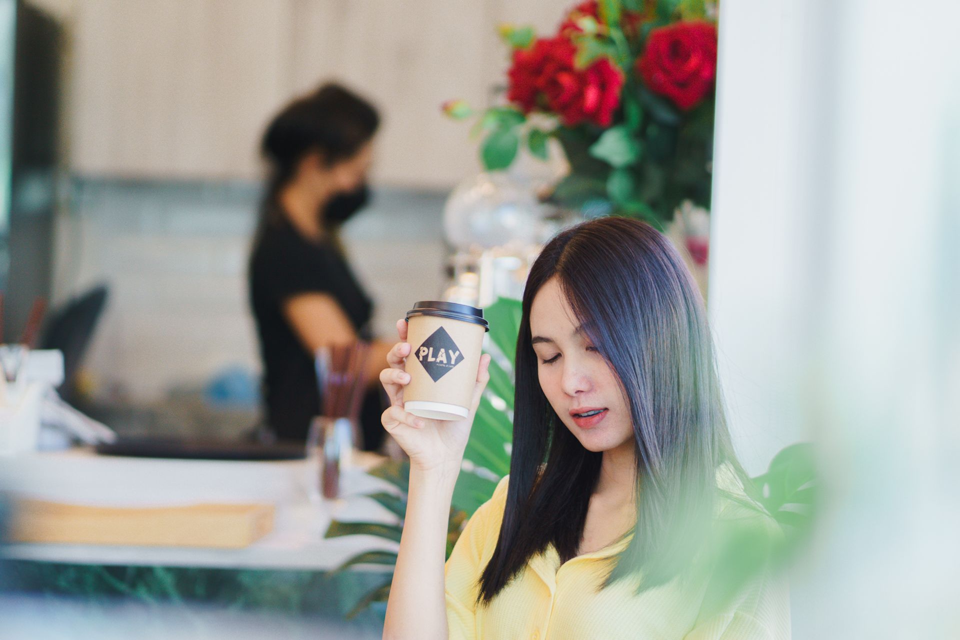 Woman holding a coffee cup, looking down in a cafe setting with a server in the background.