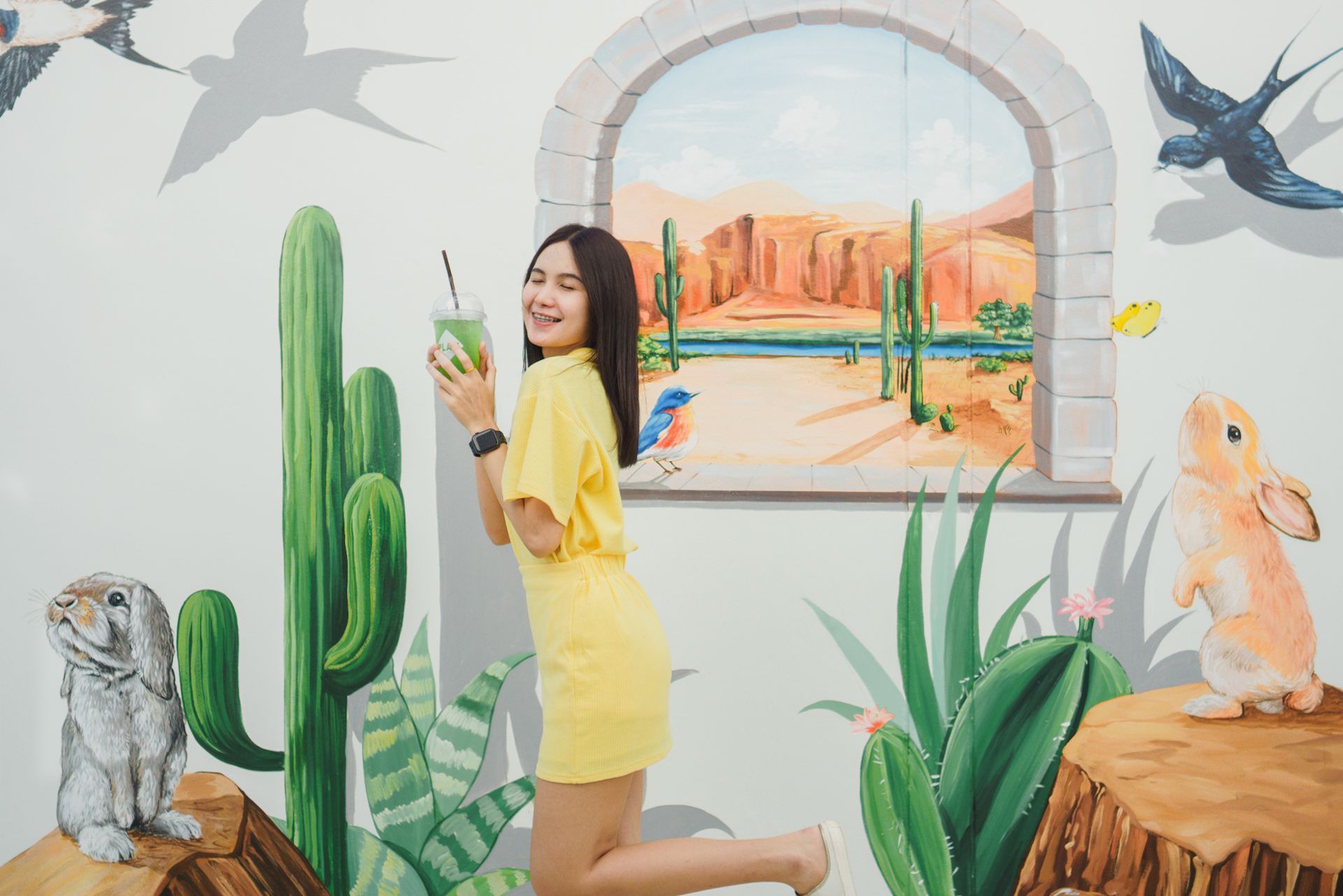 Woman in yellow dress poses with a drink in front of a desert mural.
