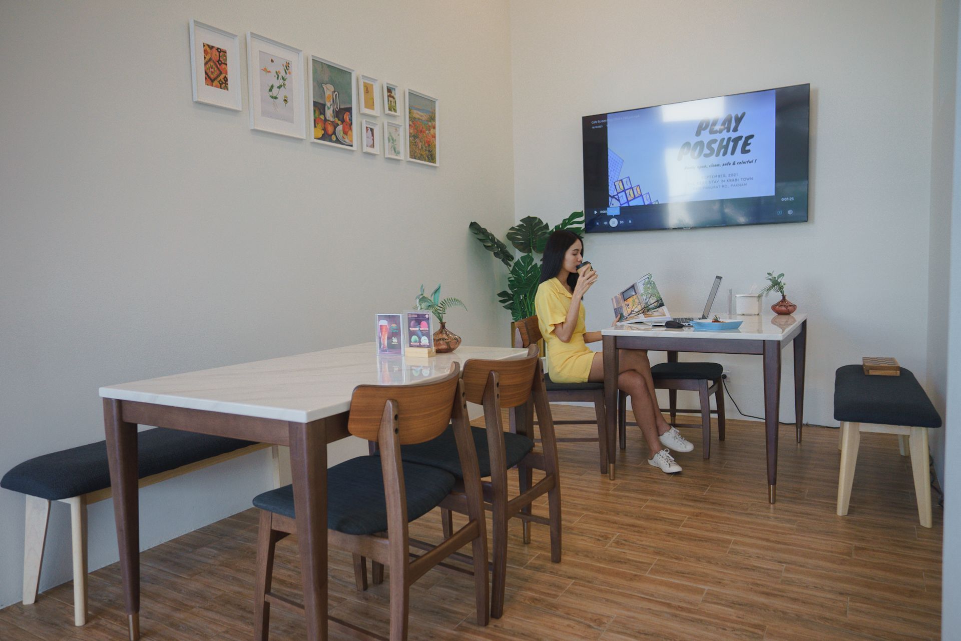 Woman in yellow dress works at a table in a cafe, looking at a laptop, with a TV in the background.