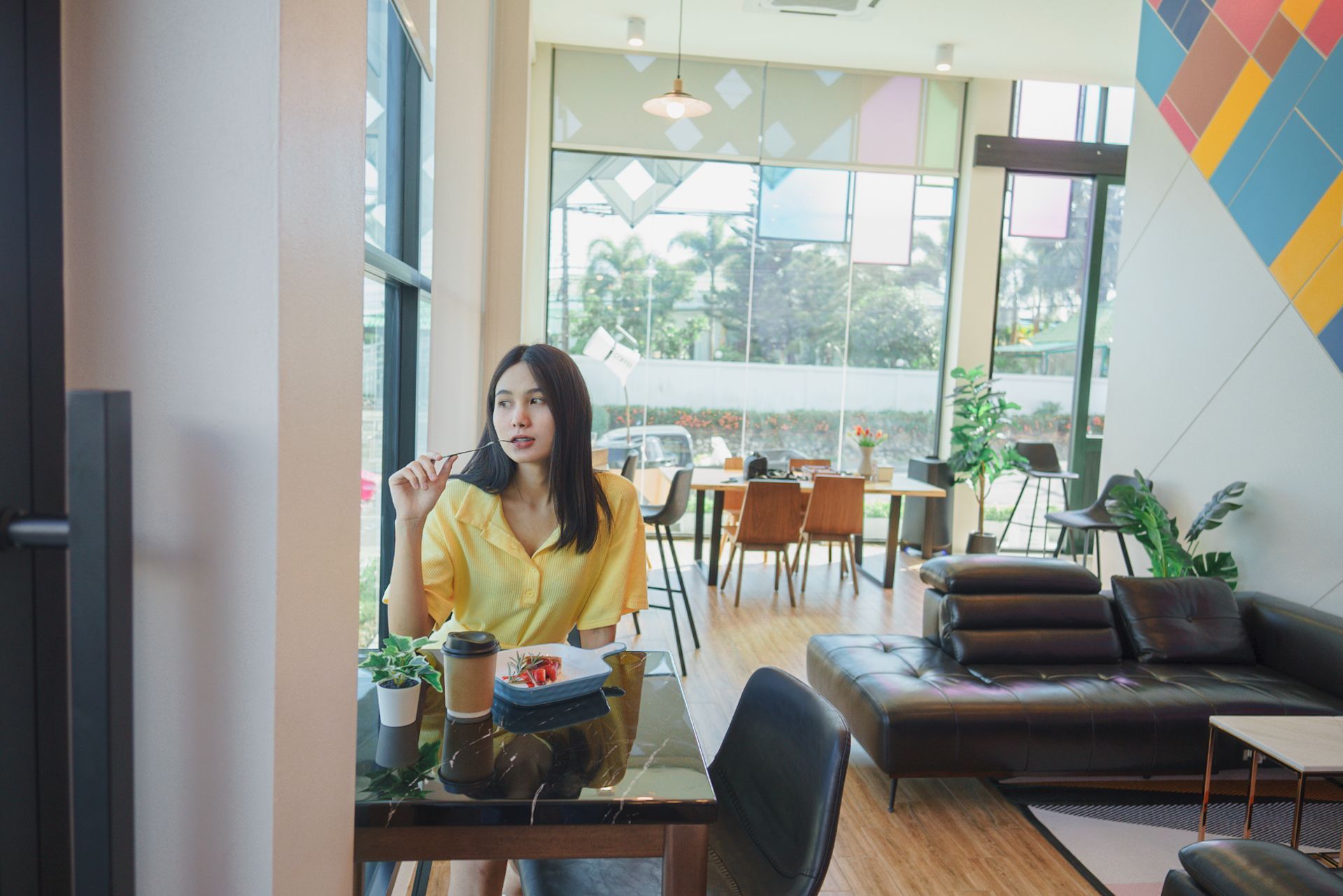 Woman in yellow shirt sits at table in cafe, looking out window. Coffee and food visible.