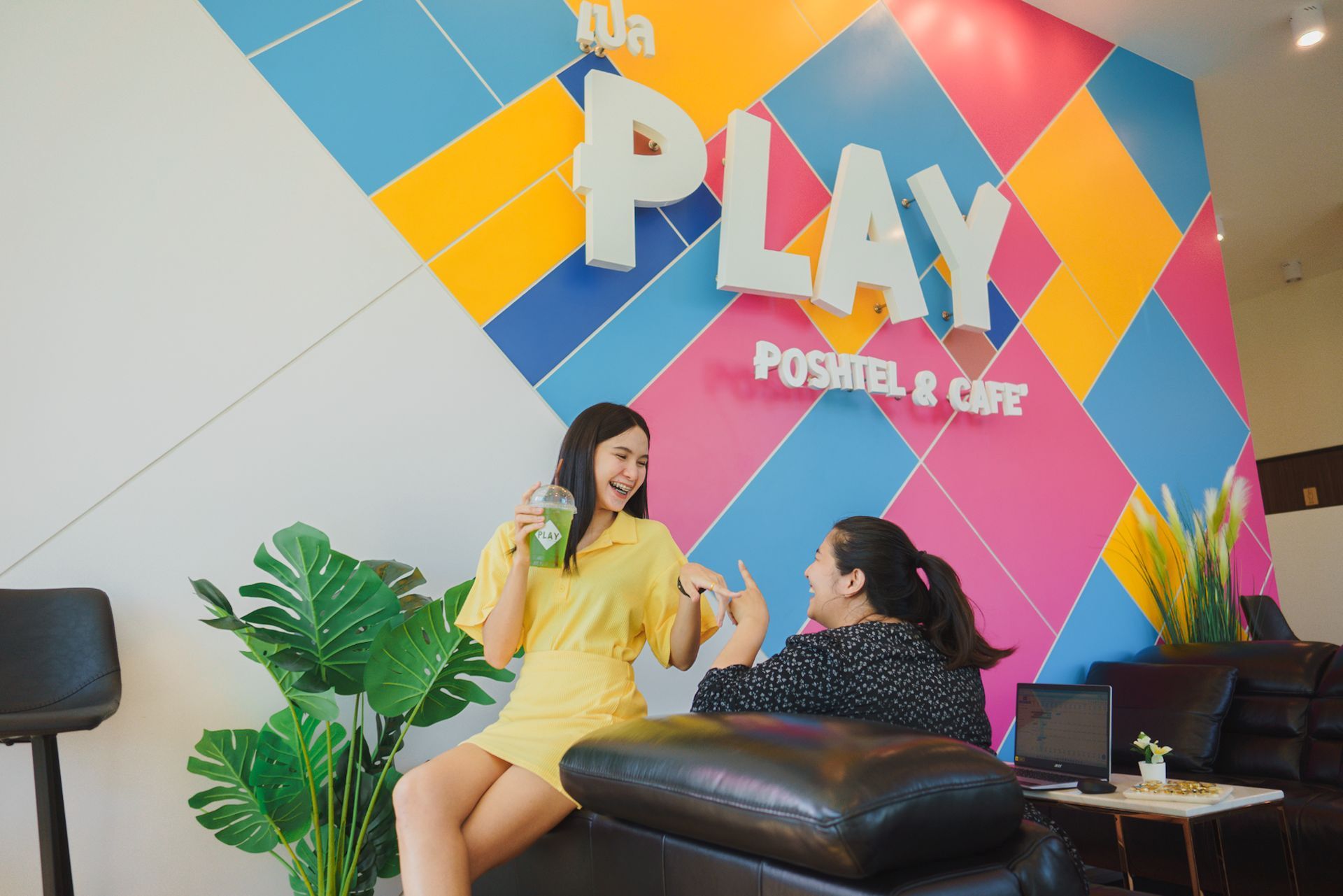 Two women laughing in a colorful cafe; one holds a green drink, the other points.