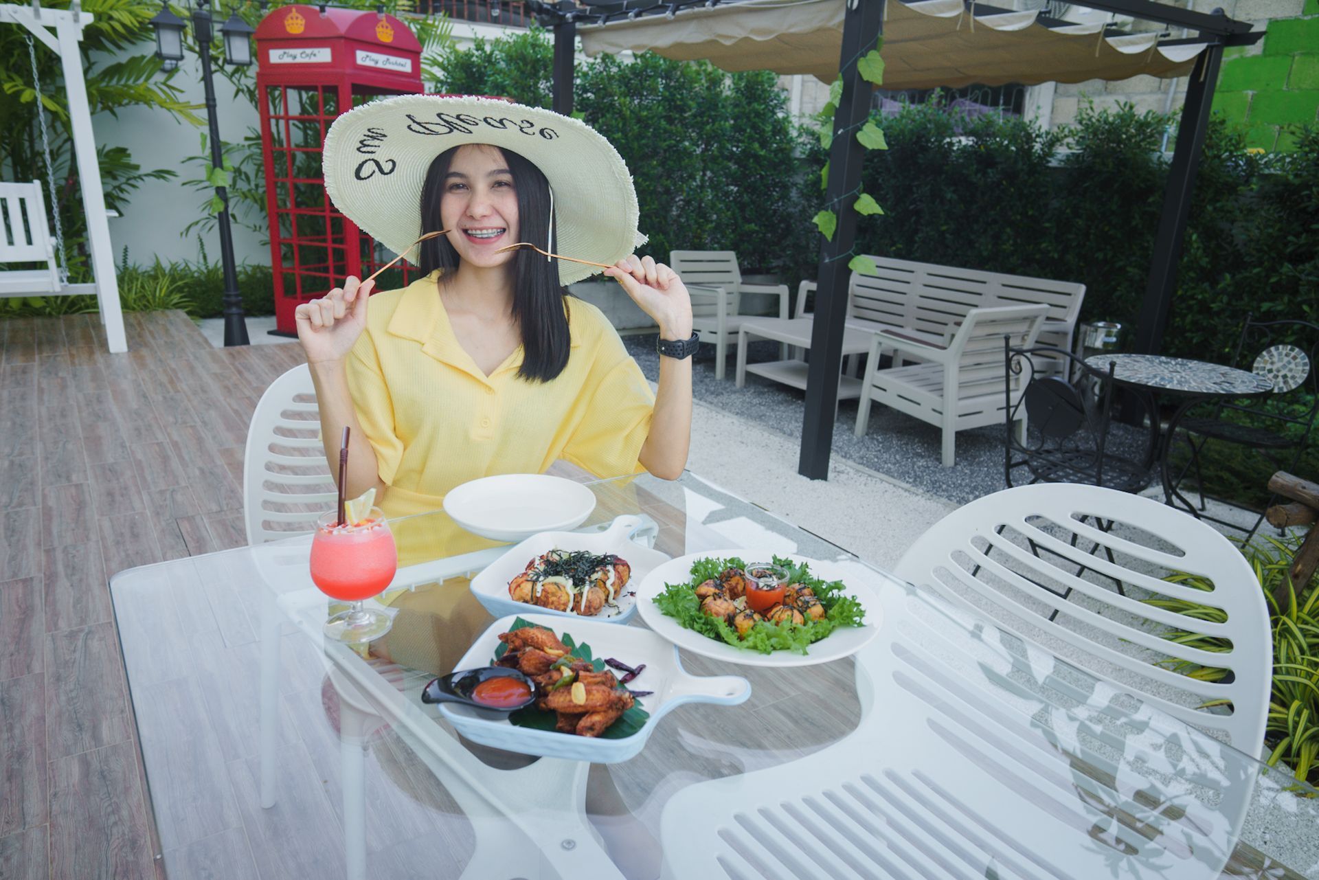 Woman in yellow dress and hat smiles at outdoor table with food and drinks.