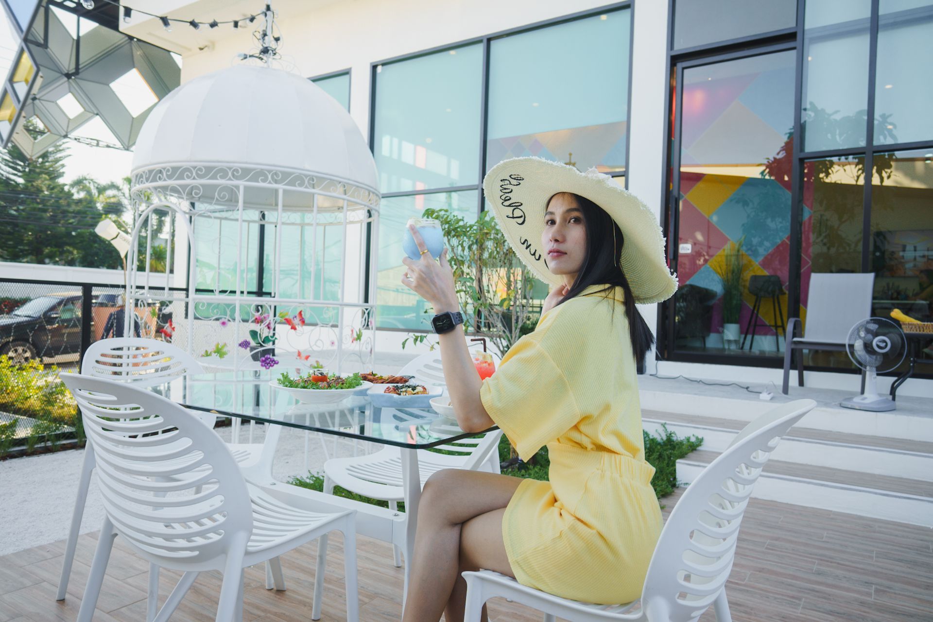 Woman in yellow dress and hat sits at outdoor table.