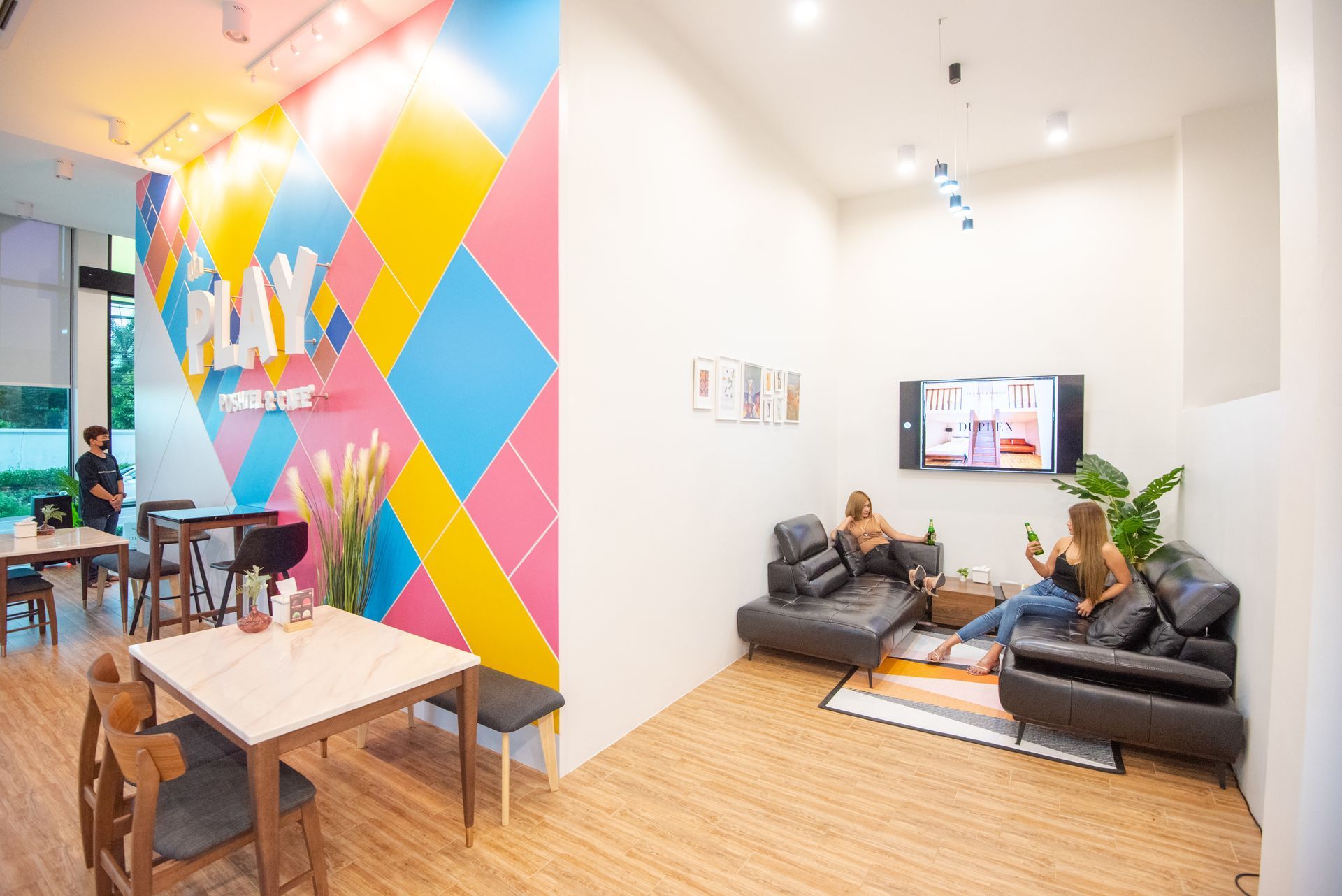 A colorful cafe with seating areas. Two people on a couch near a TV. Others at tables. 