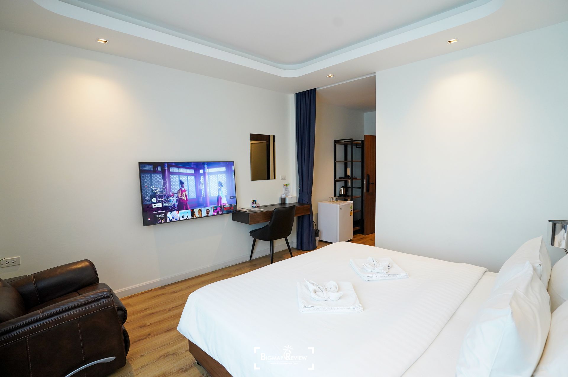 Hotel room with bed, TV, desk, chair, and a leather armchair. White walls, wooden floor, and blue curtain.