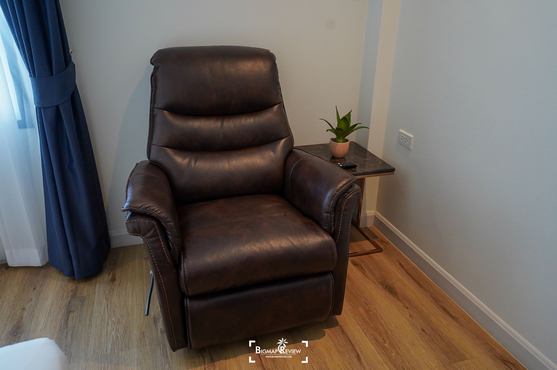Brown leather recliner chair next to a small side table with a plant, beside a curtain.