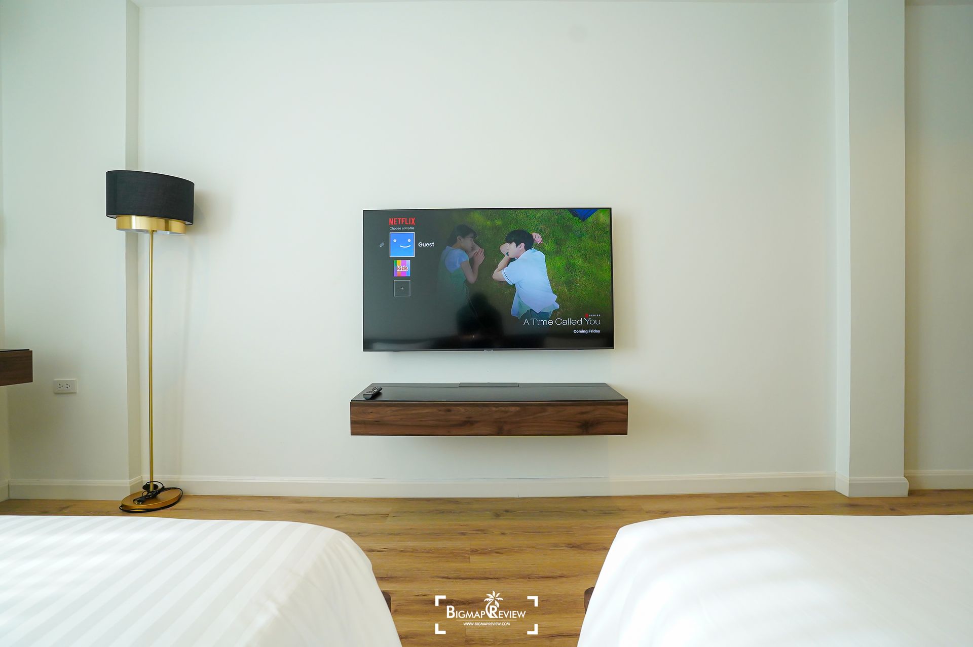 A TV on a wooden shelf above two beds in a white room, with a tall lamp to the left.