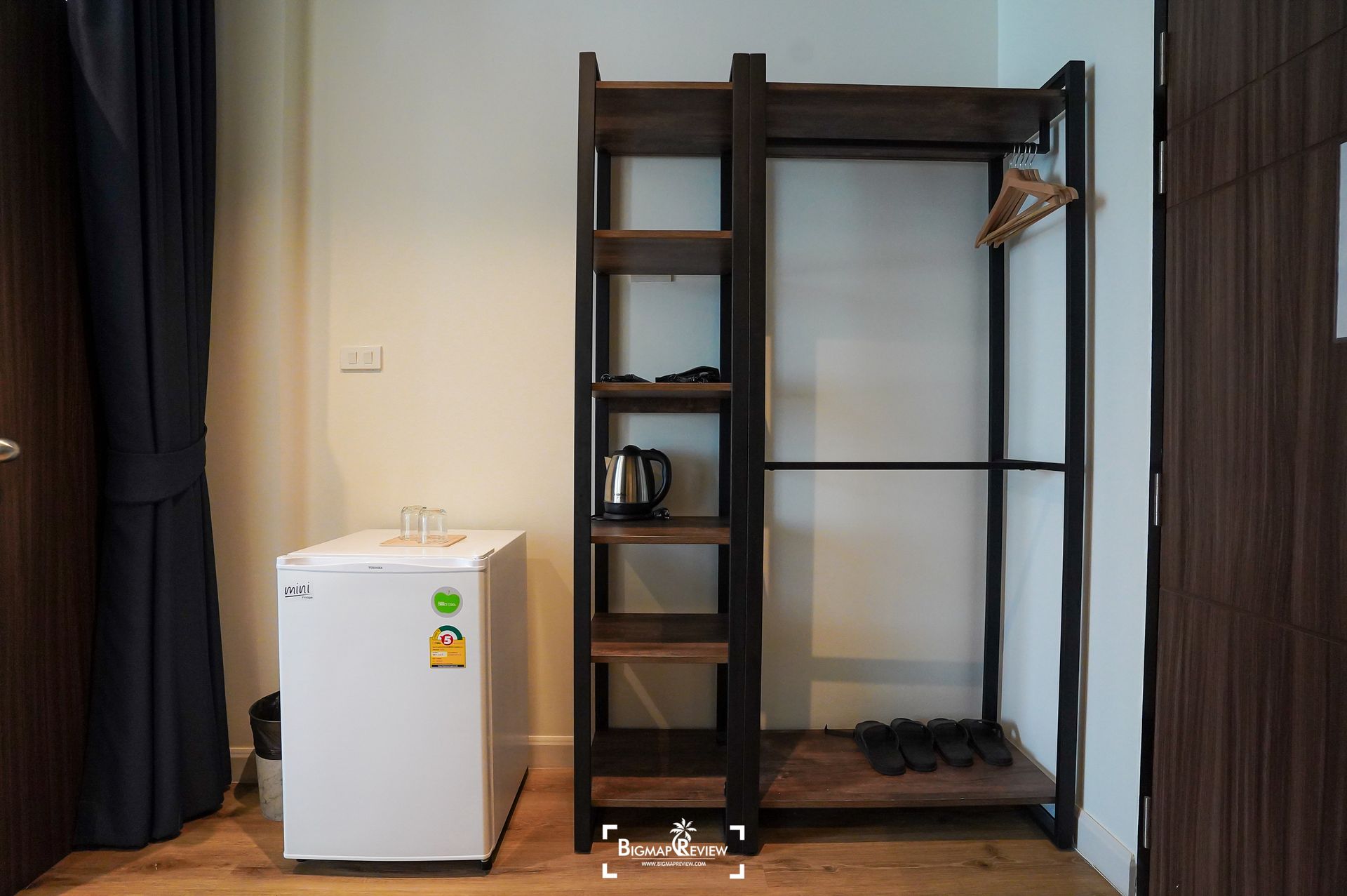 Room interior with open wardrobe, mini-fridge, and electric kettle against a white wall and wooden floor.
