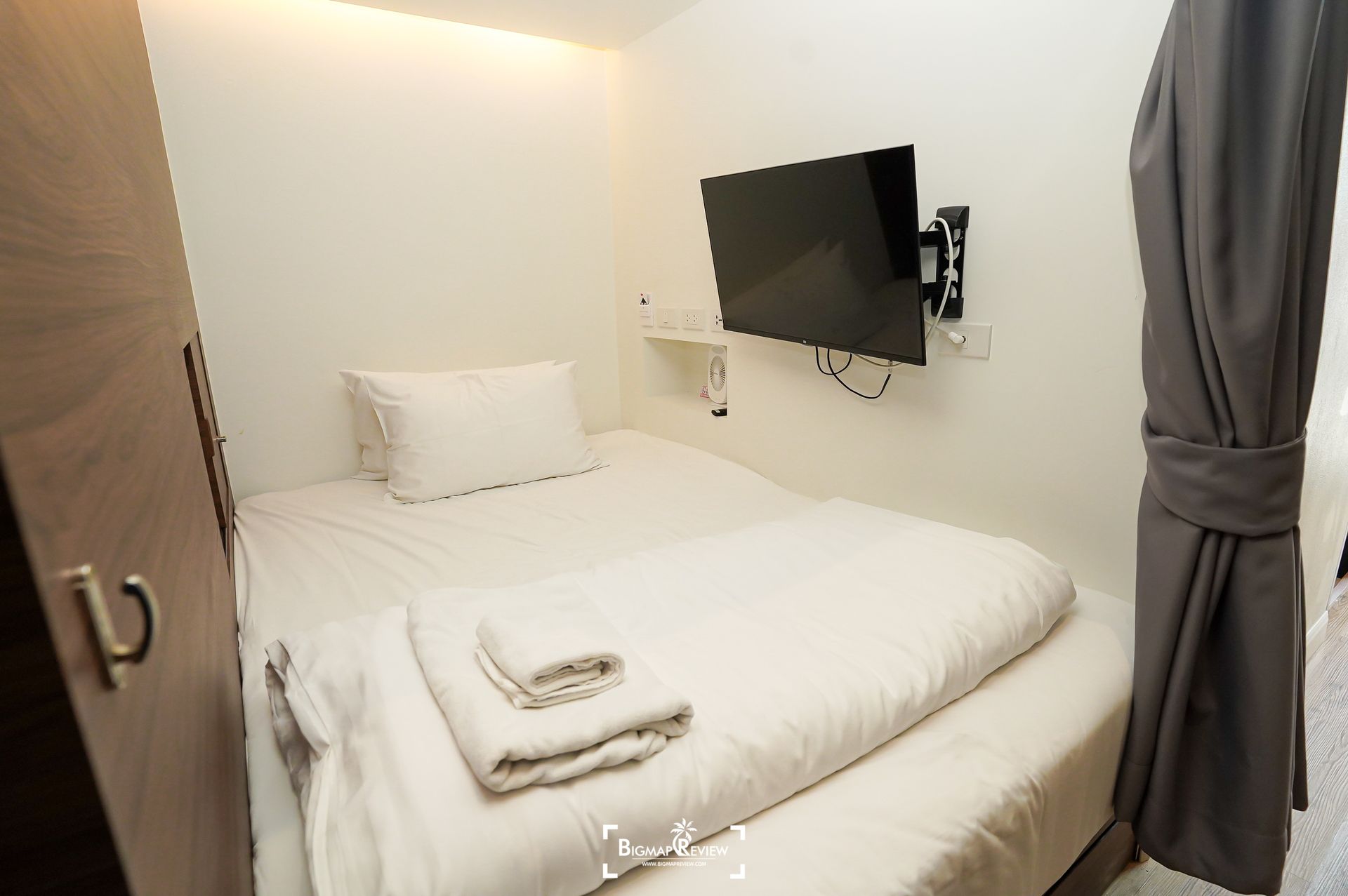 Small, white bedroom with bed, TV, and wardrobe; gray curtains hang on the right.