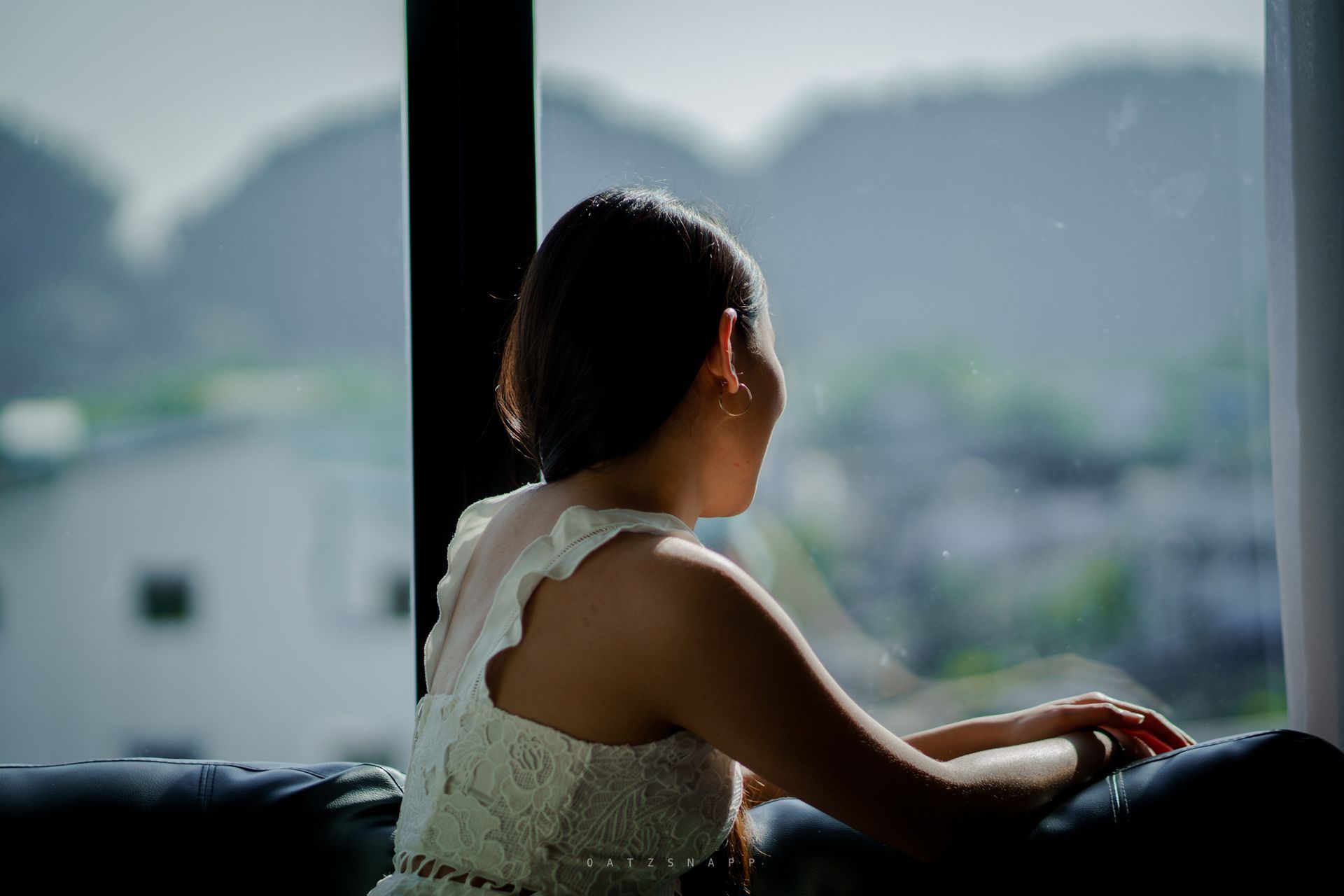 Woman looking out a window at a blurred, overcast outdoor scene; seated on a dark couch.