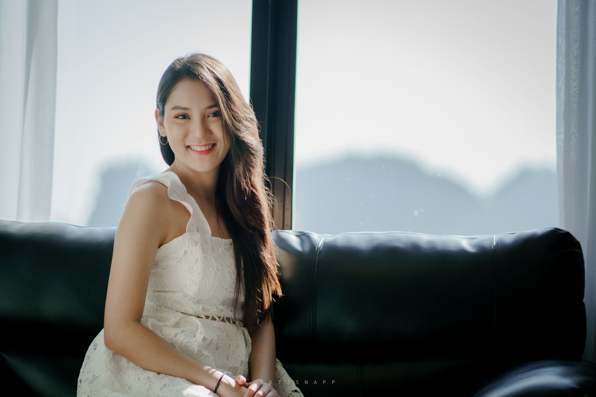 Woman in a white dress smiles while sitting on a black couch near a window. Sunlight illuminates the scene.