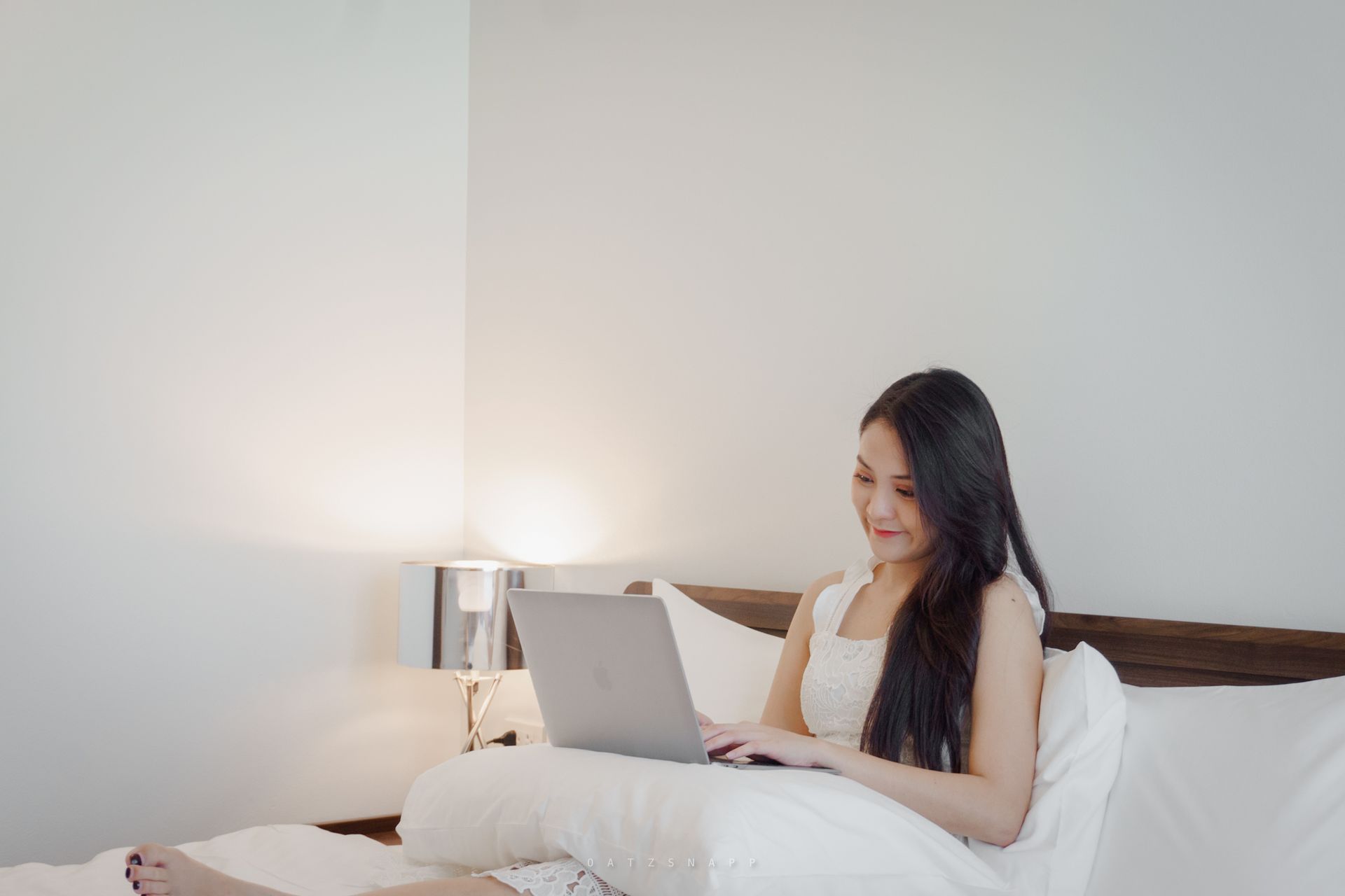 Woman sitting on a bed, using a laptop. She's smiling, near a lamp.