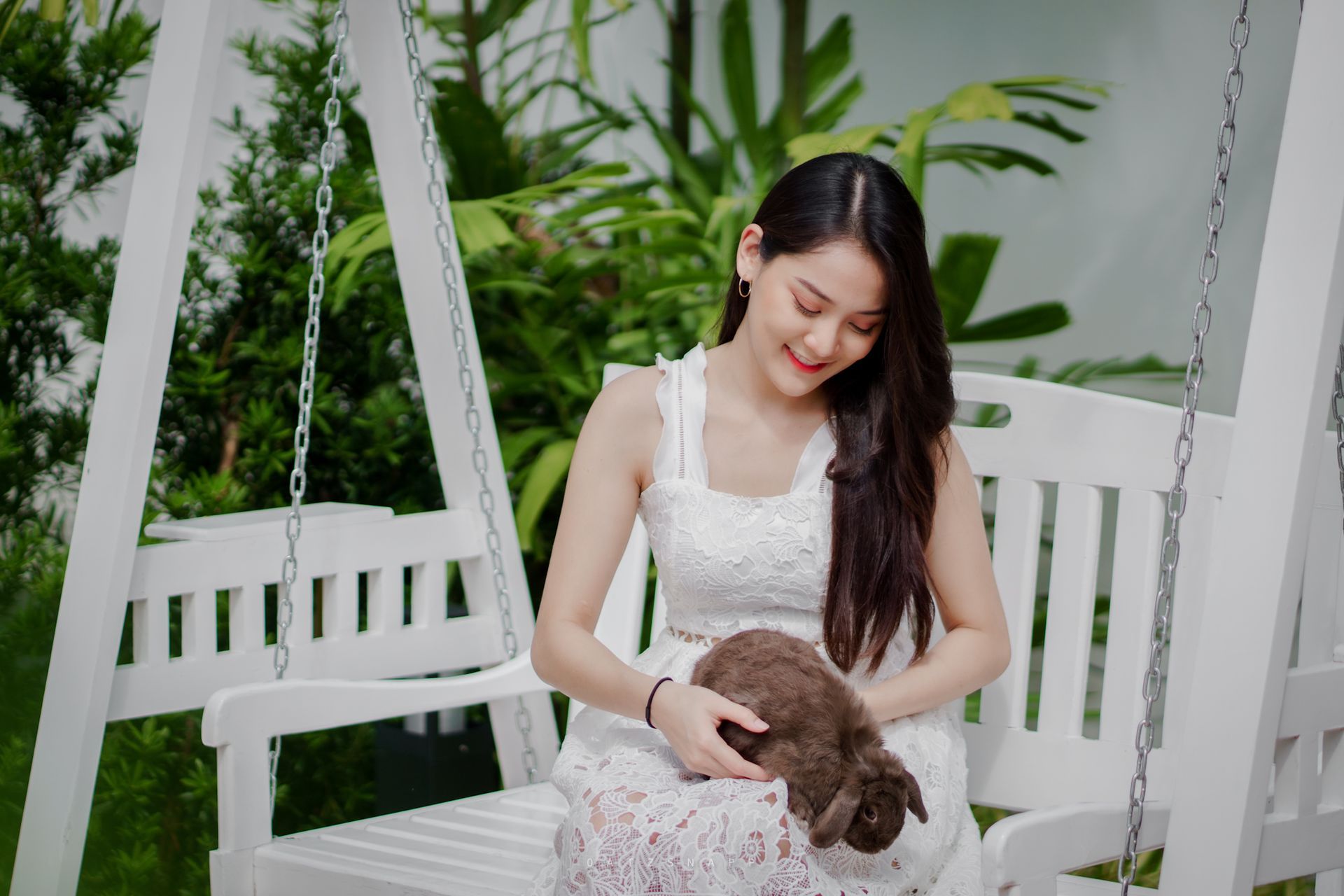 Woman in white dress smiles, petting a brown rabbit on a white swing outdoors.
