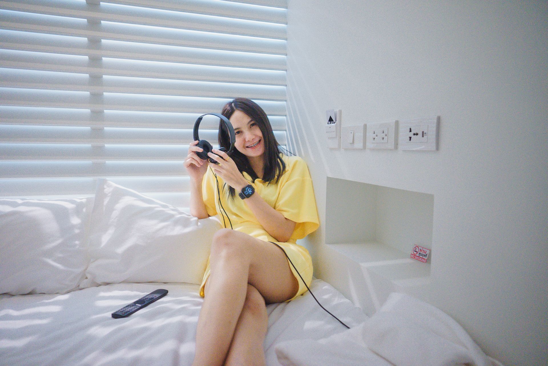 Woman in yellow dress sits on bed, holding headphones, smiling. White room, blinds, and outlets visible.