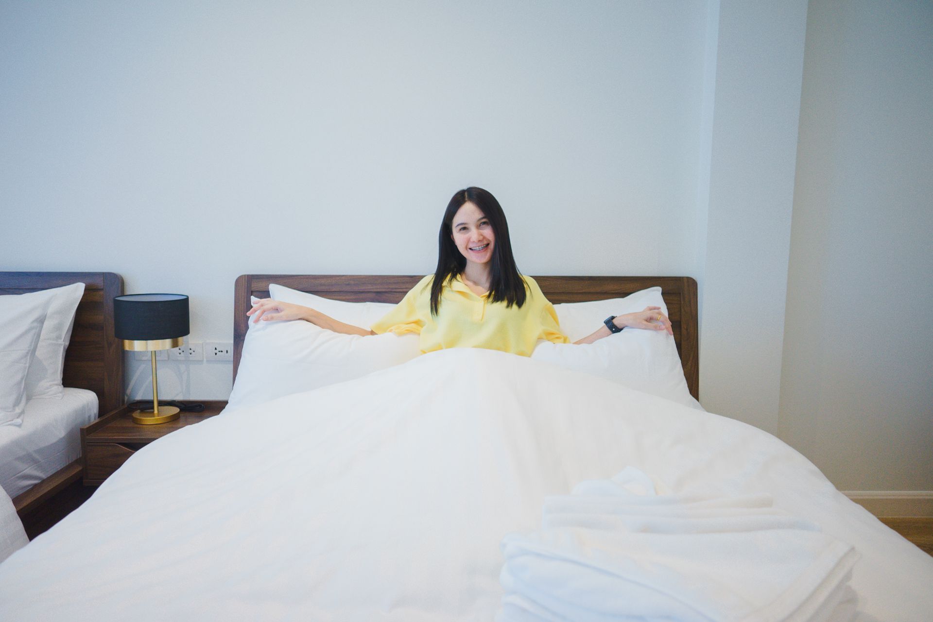 Woman in yellow shirt sits on a large white bed, arms outstretched, smiling.