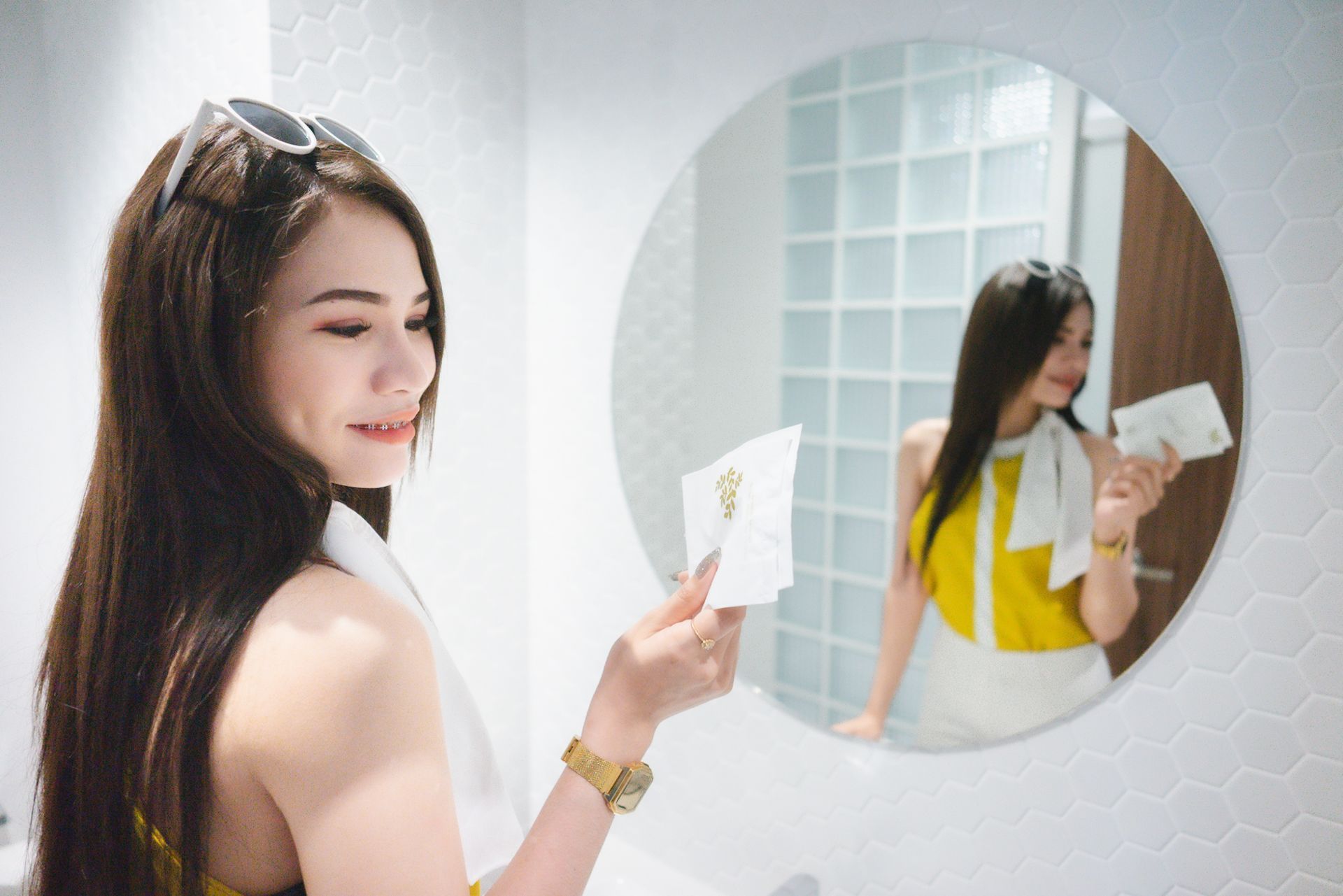 Woman in yellow top and white skirt holding tissues in bathroom, looking at mirror.