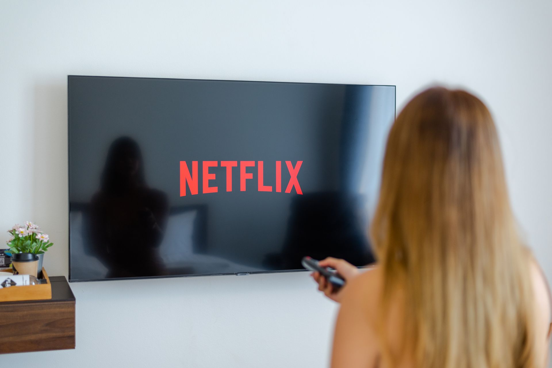 Person with long blonde hair holding a remote, facing a TV displaying the Netflix logo.
