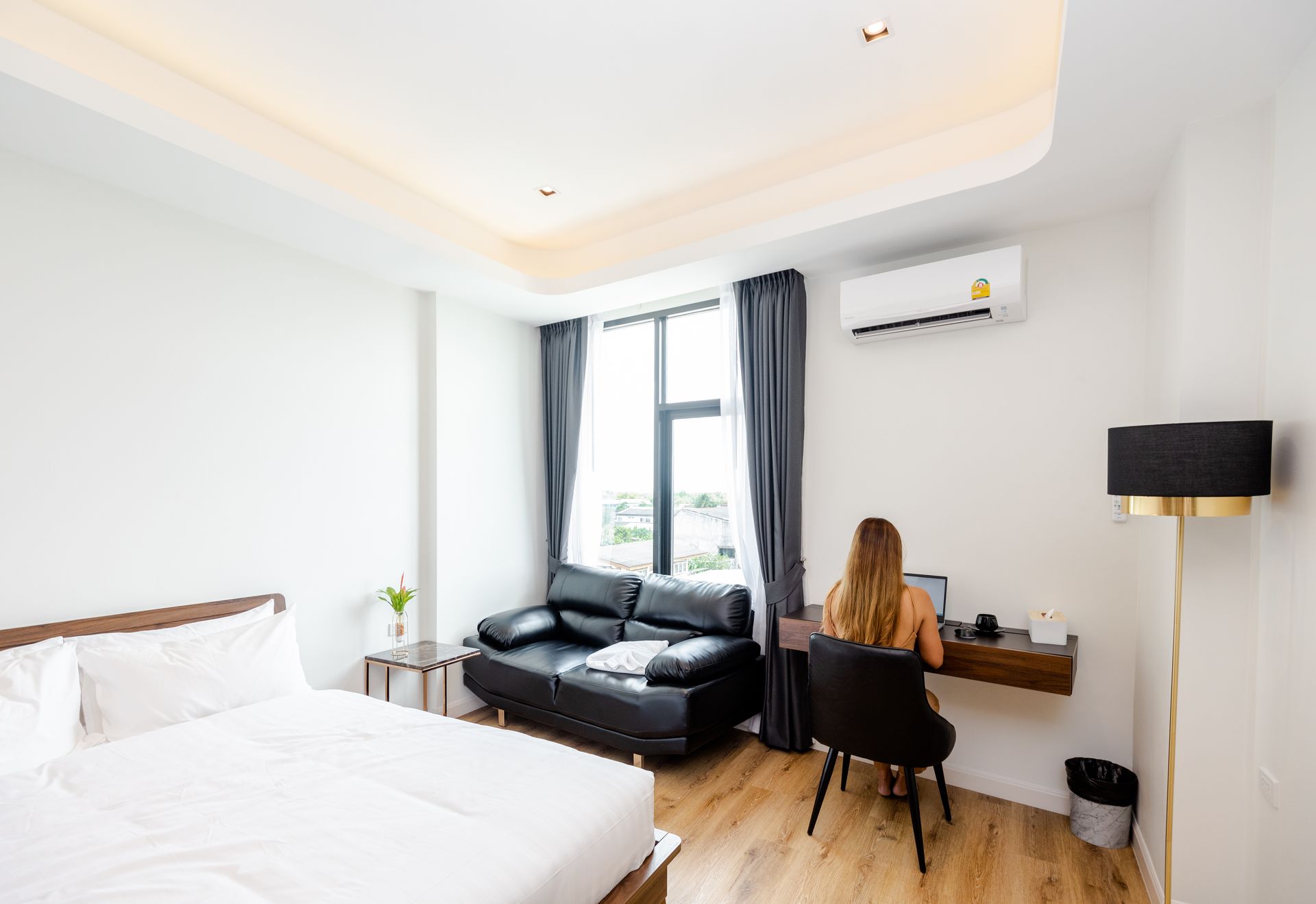 Woman working on laptop at desk in a bright hotel room with a bed and a black leather sofa.