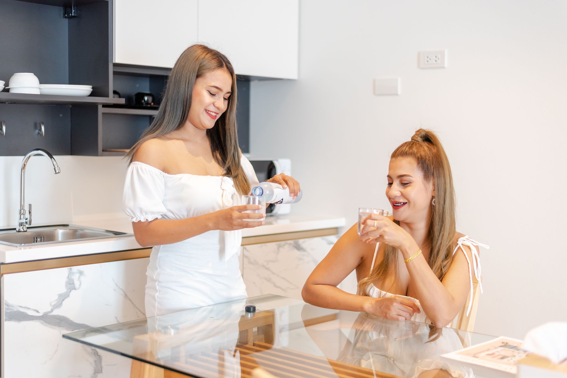 Two women in a kitchen: one pours water into a glass while the other sits, smiling.