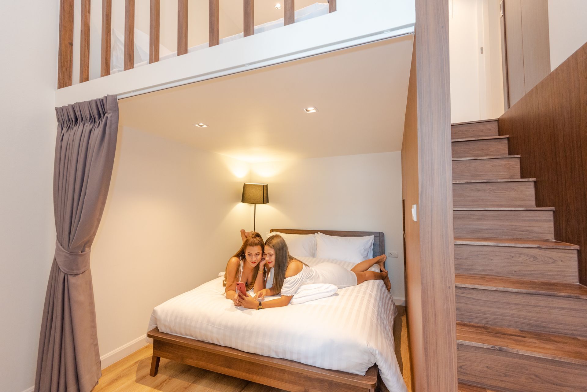 Two people in bed under a loft, smiling. Wooden stairs to the right, curtains on the left.