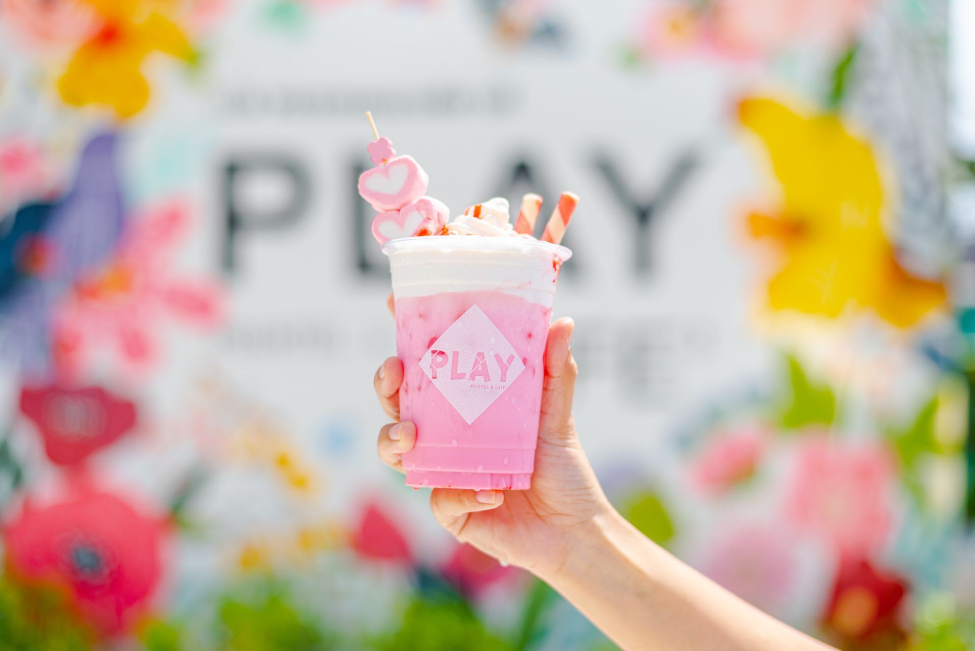 A hand holding a pink drink with marshmallows, whipped cream, and a straw, in front of a colorful floral backdrop.