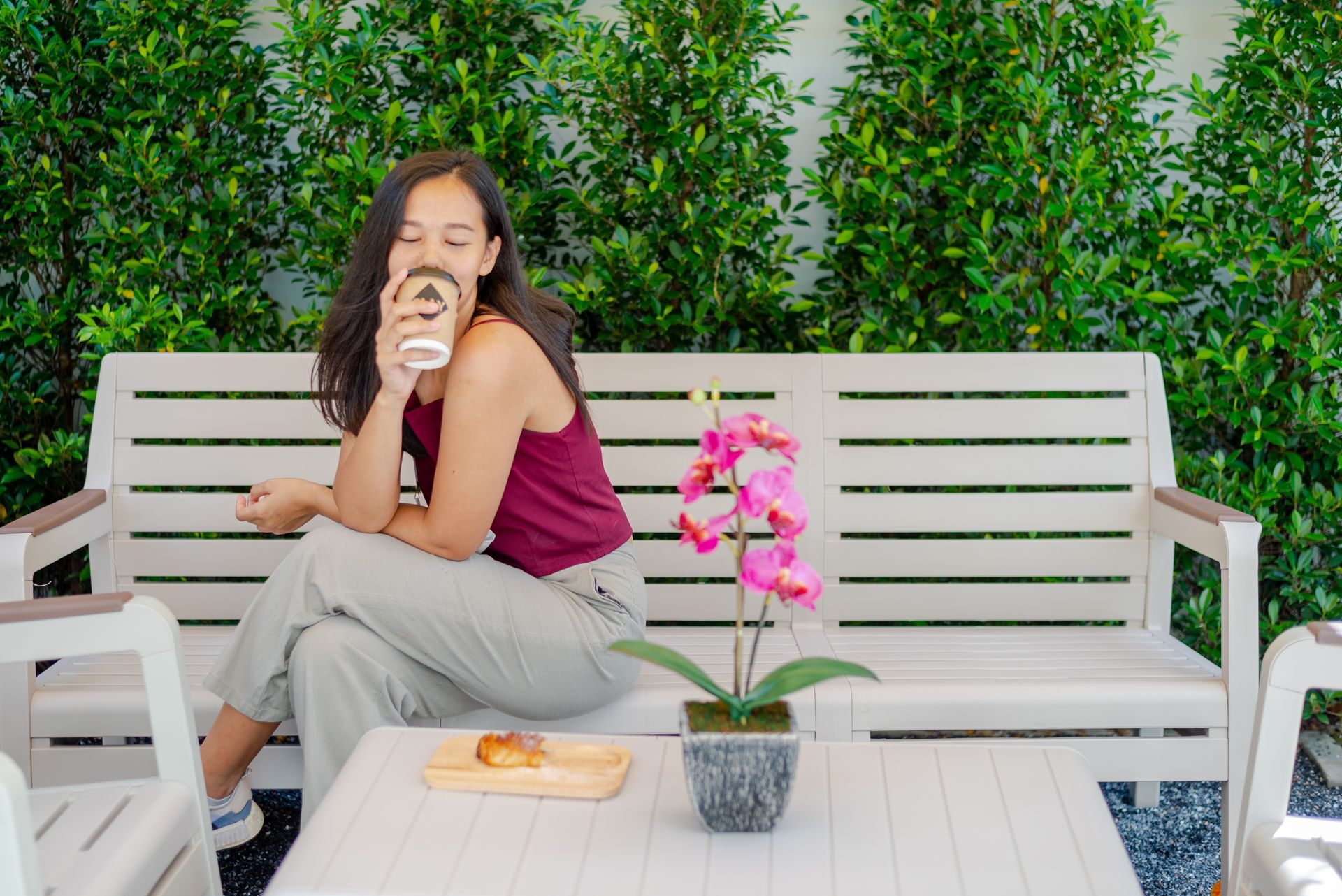 Woman sitting on white bench, enjoying coffee, next to pink orchid, in front of green hedge.