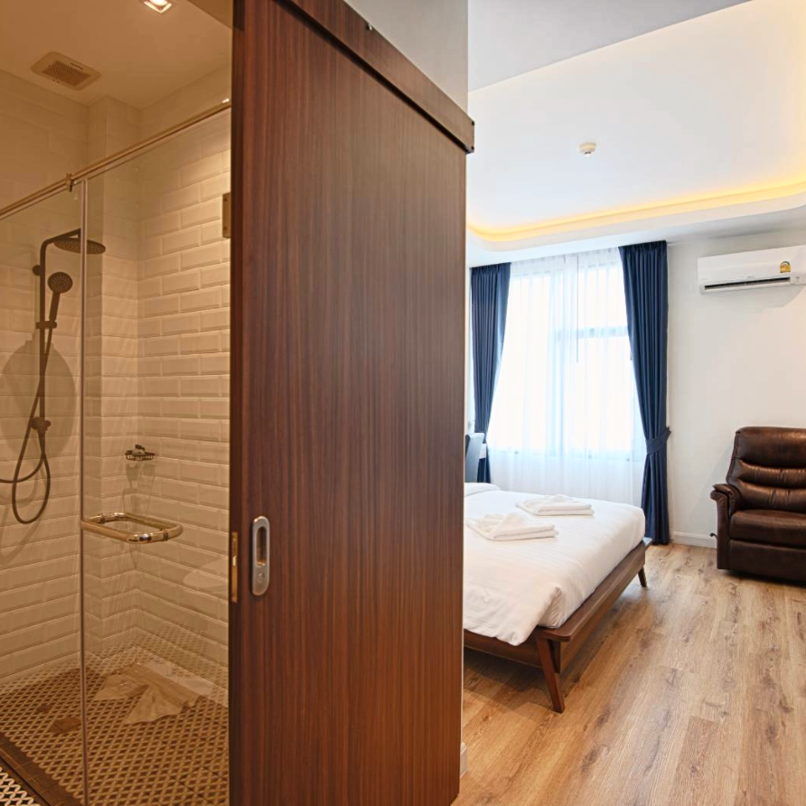 Bathroom shower with glass door and bedroom with bed and recliner visible.