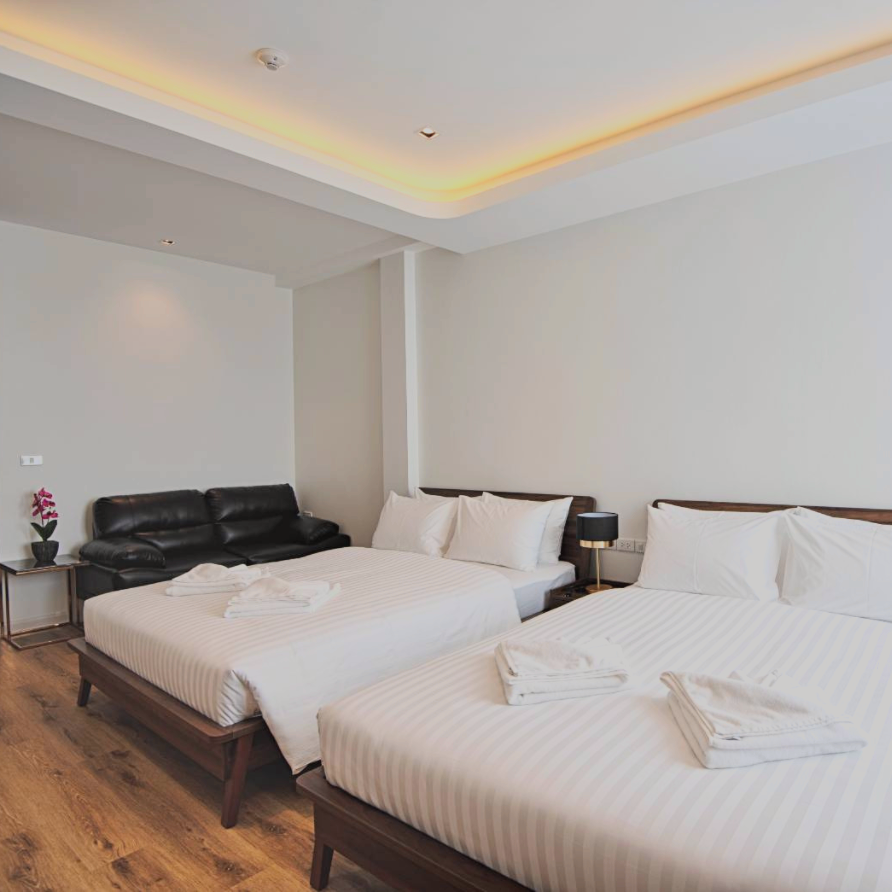 Hotel room with two beds, a black sofa, and a table. White walls, wooden floors, and ceiling lighting.