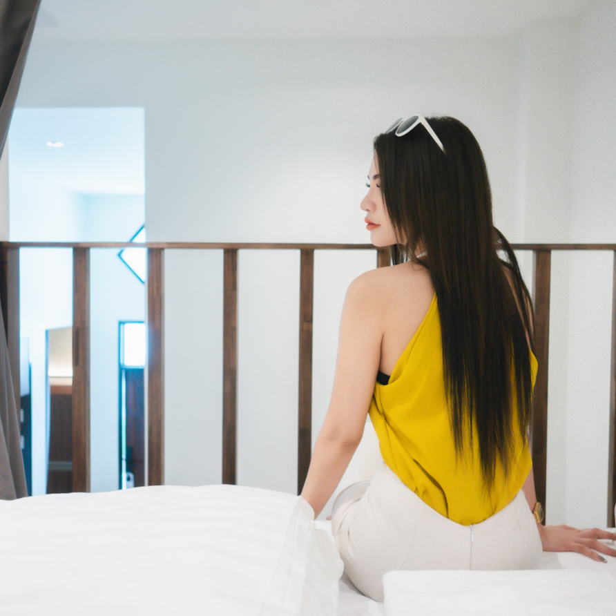 Woman sitting on a white bed, wearing a yellow top and white pants, looking away.