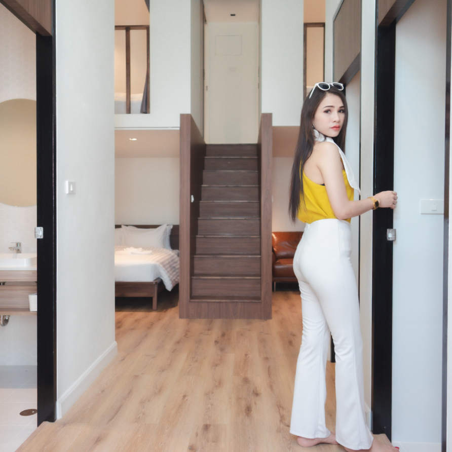 Woman in white pants and yellow top stands in a modern hallway with stairs, doorway, and bedroom.