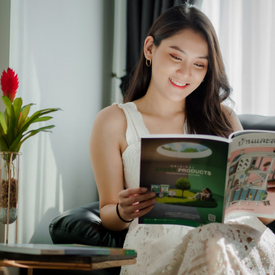 Woman smiling, reading a catalog with green cover, sitting indoors near a window.