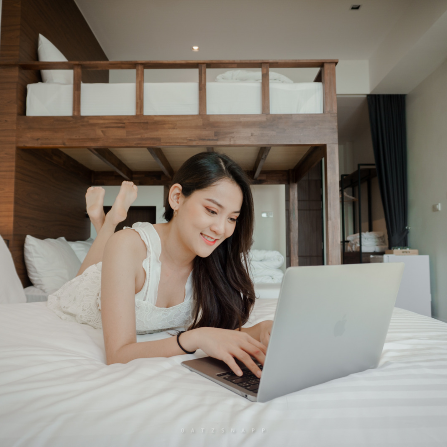 Woman lying on bed using a laptop in a hotel room, smiling.