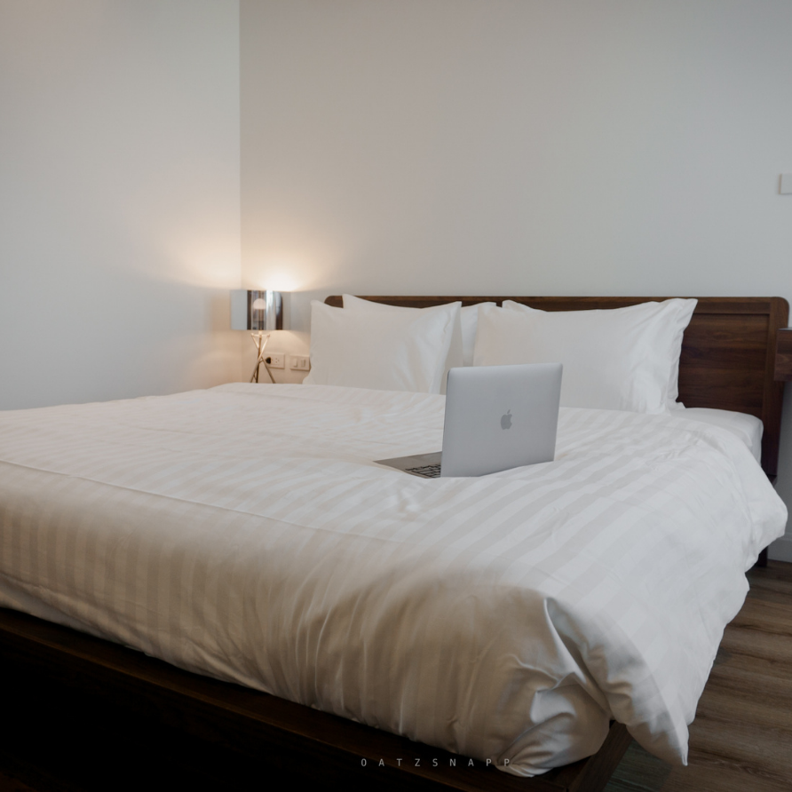 Bed with white linens, wooden headboard, and laptop. Room has white walls and a reading light.