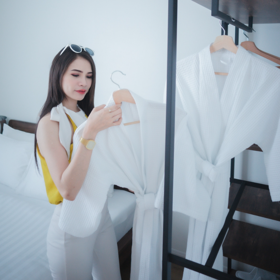 Woman in white pants and yellow top choosing a white robe in a hotel room.