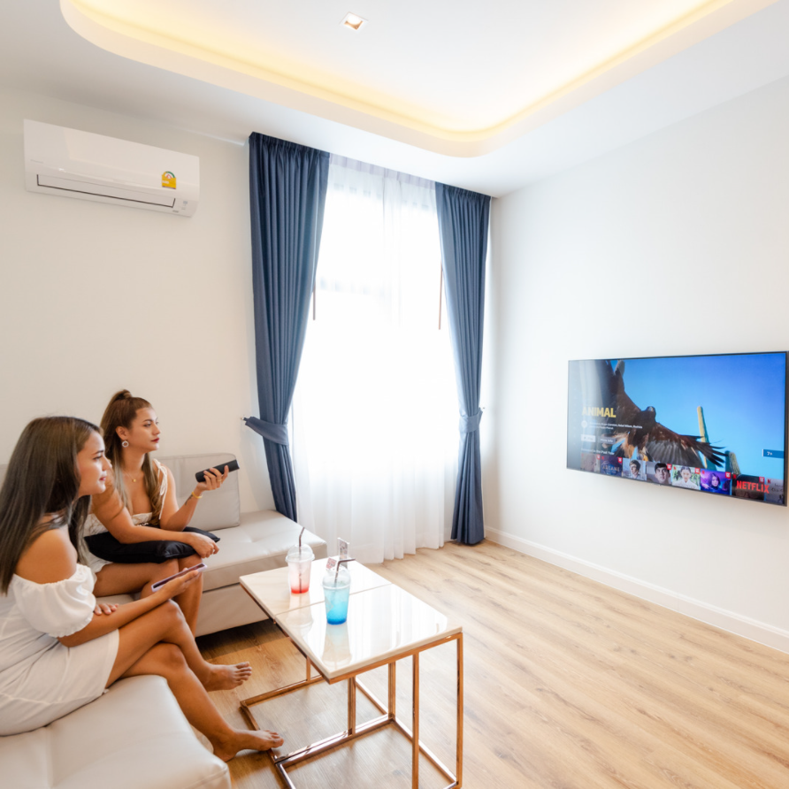 Two women sit on a couch watching TV in a bright room with blue curtains and wooden floor.