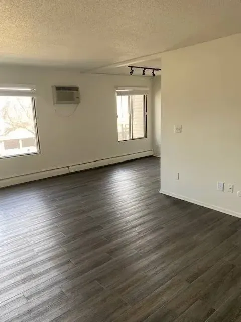 Empty room with dark wood-look flooring, white walls, windows, and an air conditioner.