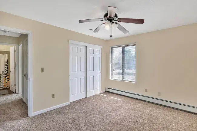 Bedroom with tan walls, carpet, white closet doors, window, and ceiling fan.