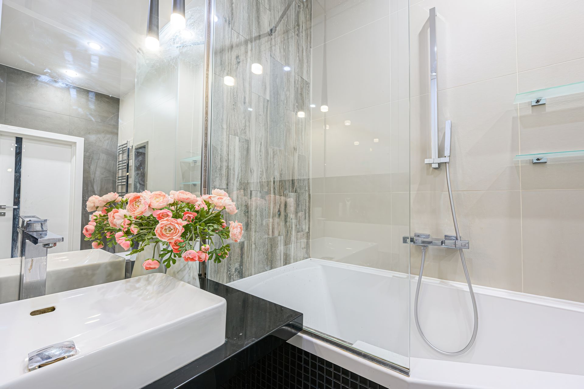 A modern, neutral-toned bathroom featuring a sleek white sink with flowers and a bathtub behind a glass partition.