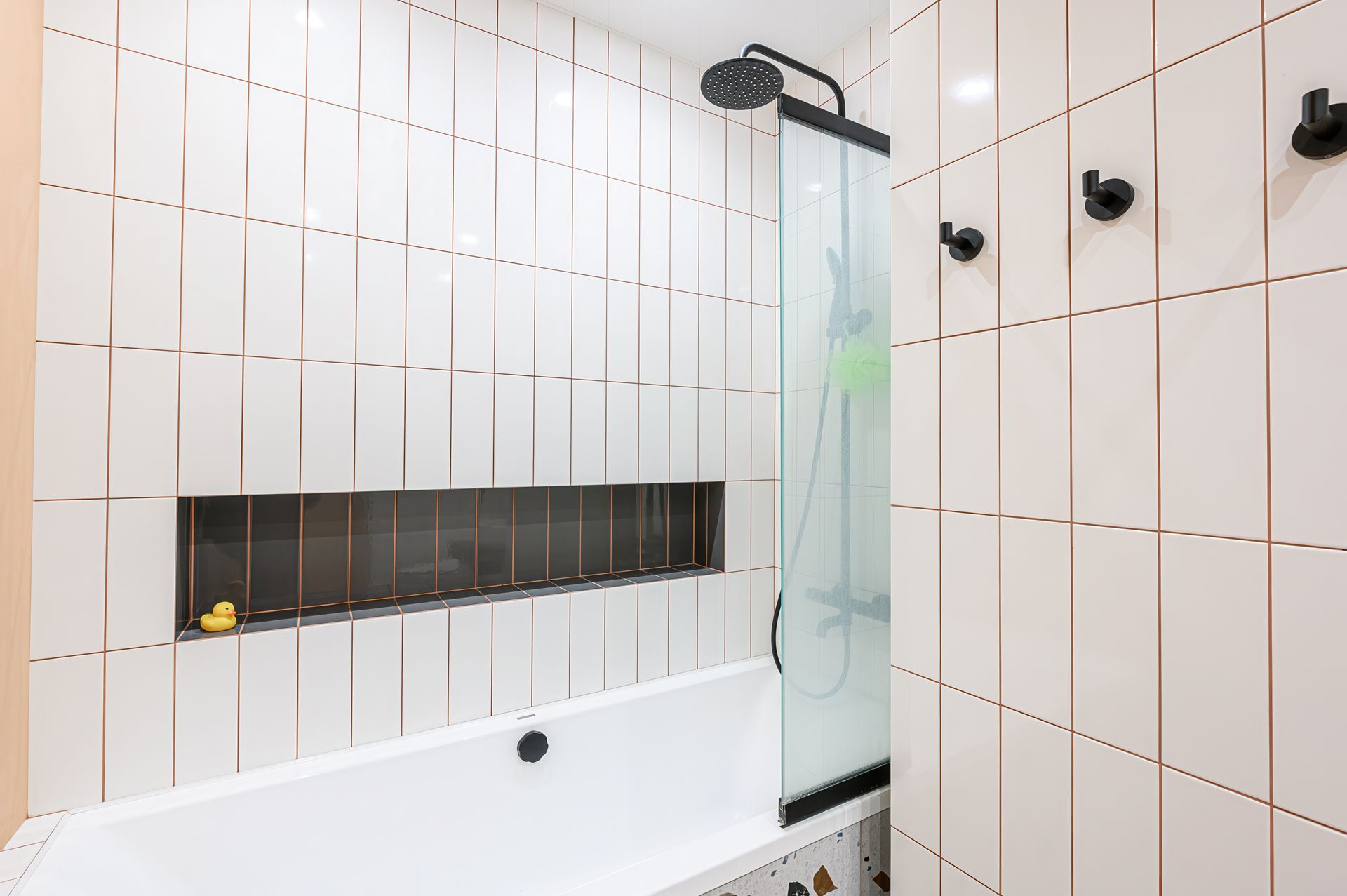 A white bathtub with rose gold grout lines, a shower head, glass screen, recessed shelf, and three black wall hooks.