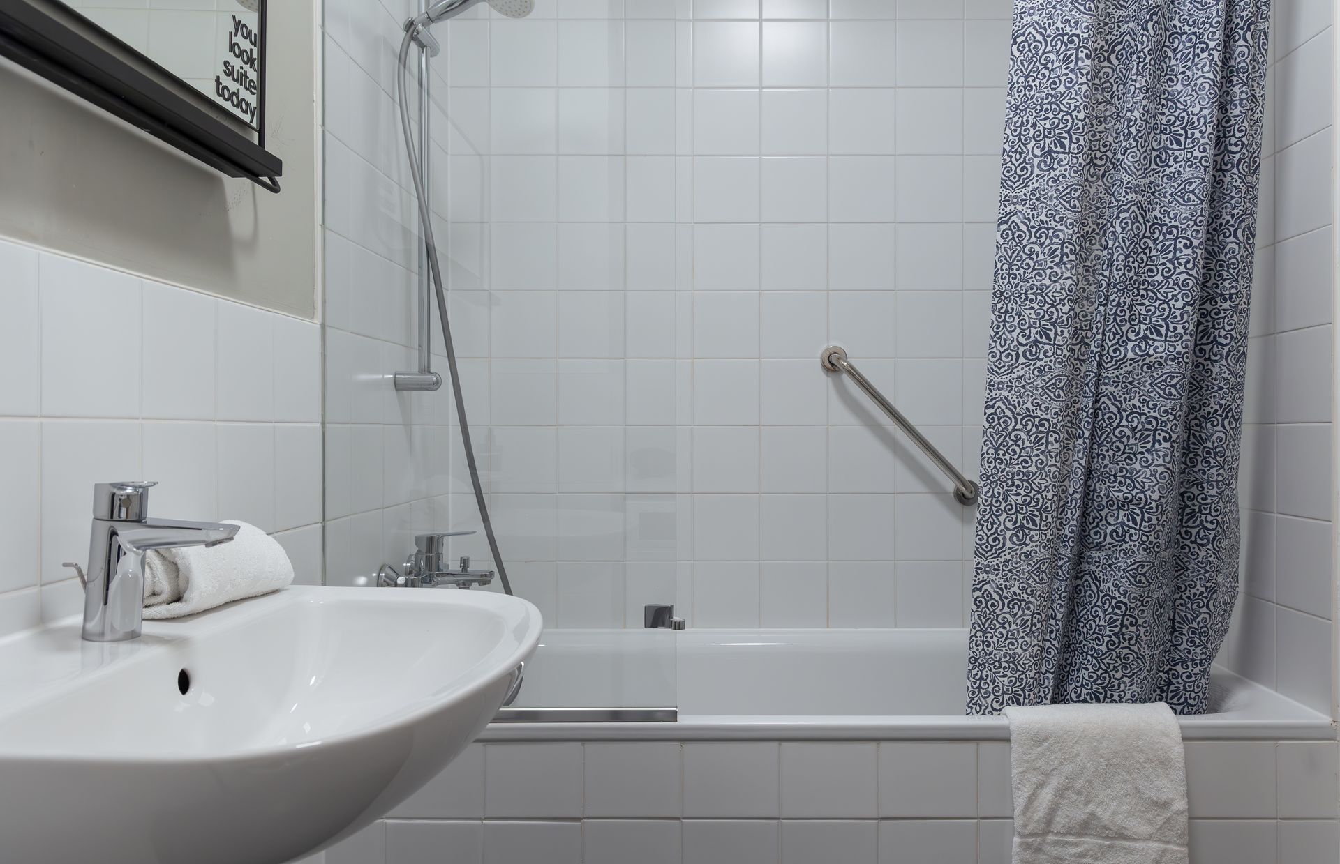 A white bathroom featuring a porcelain sink, a bathtub with a patterned shower curtain, and a wall-mounted grab bar.