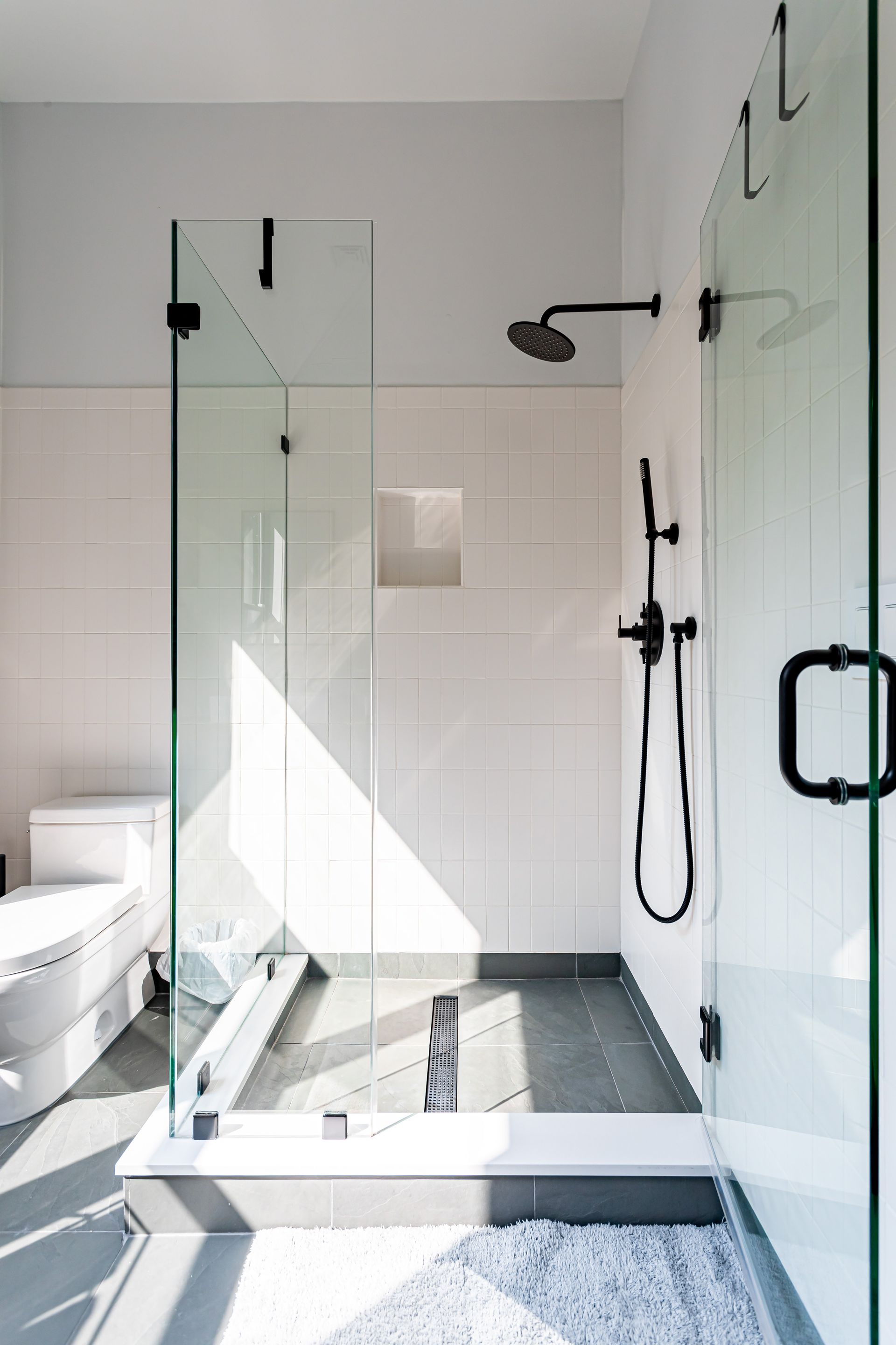 A glass-enclosed shower with matte black fixtures, white tiled walls, and a dark grey tiled floor next to a white toilet.